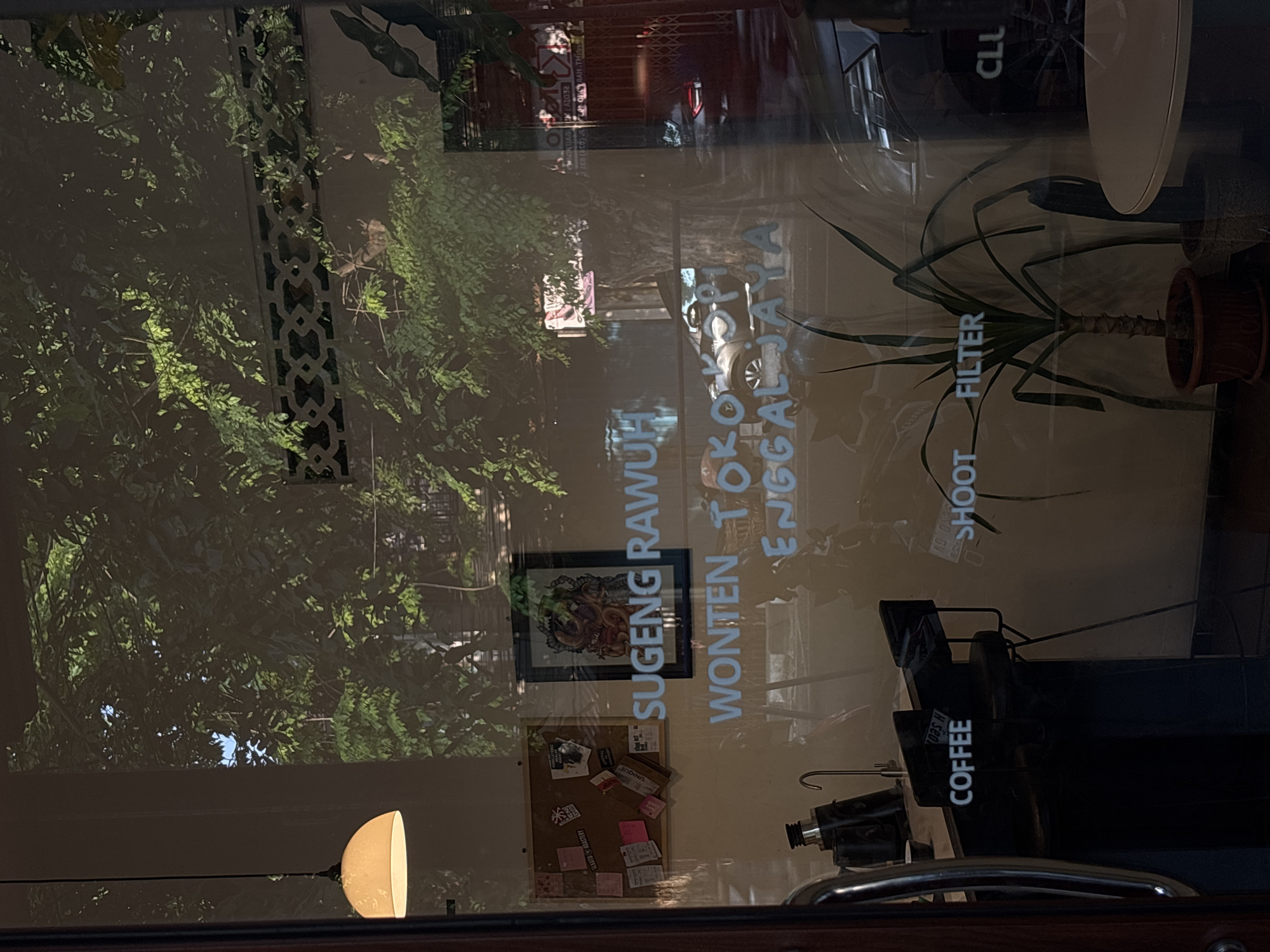 This image captures a reflection on a glass storefront, likely in Semarang, Indonesia. The reflection shows lush green trees and a decorative lattice fence against a bright sky, suggesting an outdoor environment. The storefront itself features white text on the glass, which appears to be signage. Prominently displayed are the words "SUGENG RAWUH," followed by "WONTEN TOKO KOPI ENGGALJAYA." Below this, further text is visible, including "COFFEE," "SHOOT," "FILTER," and "CLU." Inside the establishment, a corkboard with various notes and a framed artwork are visible. A light fixture hangs from the ceiling. A plant is also visible inside the shop, as well as part of a table and chairs, indicating it is likely a coffee shop or cafe. The overall impression is that of a bright, possibly sunny day, with the interior of the establishment dimly lit in contrast to the external reflection.