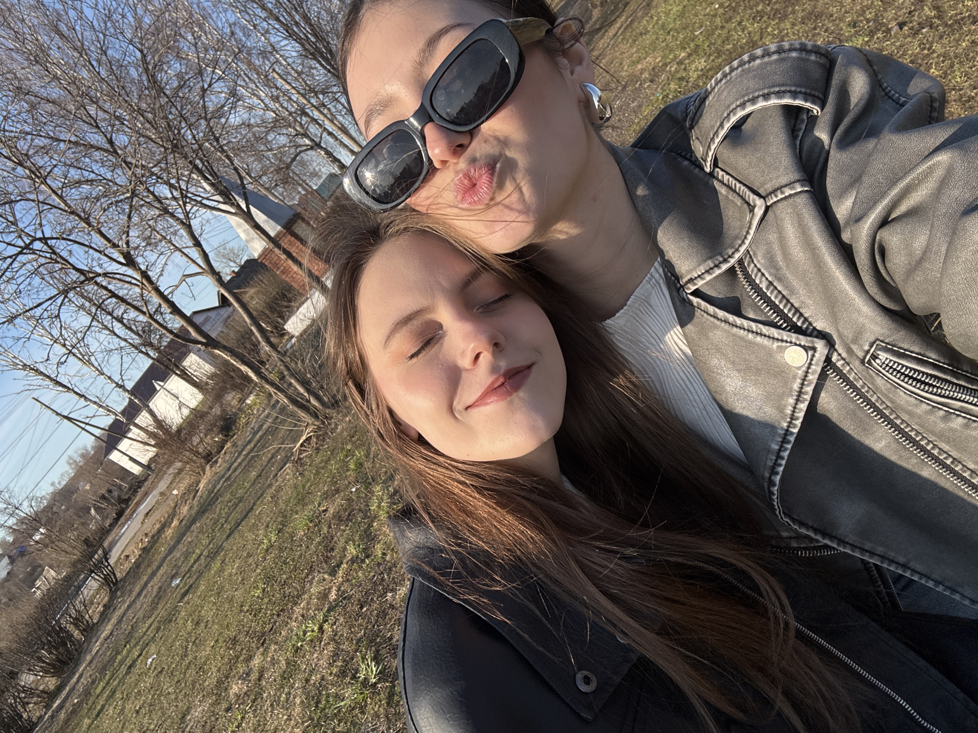 The image is a selfie of two women in an outdoor setting. The location appears to be in Фоки, Russia, considering the request.
The woman in the foreground has closed eyes and long, light brown hair. She is wearing a dark-colored jacket. The woman in the background is slightly out of focus and wearing sunglasses and a grey leather jacket. She is puckering her lips as if blowing a kiss. A silver heart-shaped earring is visible on her left ear.
The background consists of a grassy area, bare trees, and a building with a peaked roof in the distance. Power lines are also visible against the clear sky.