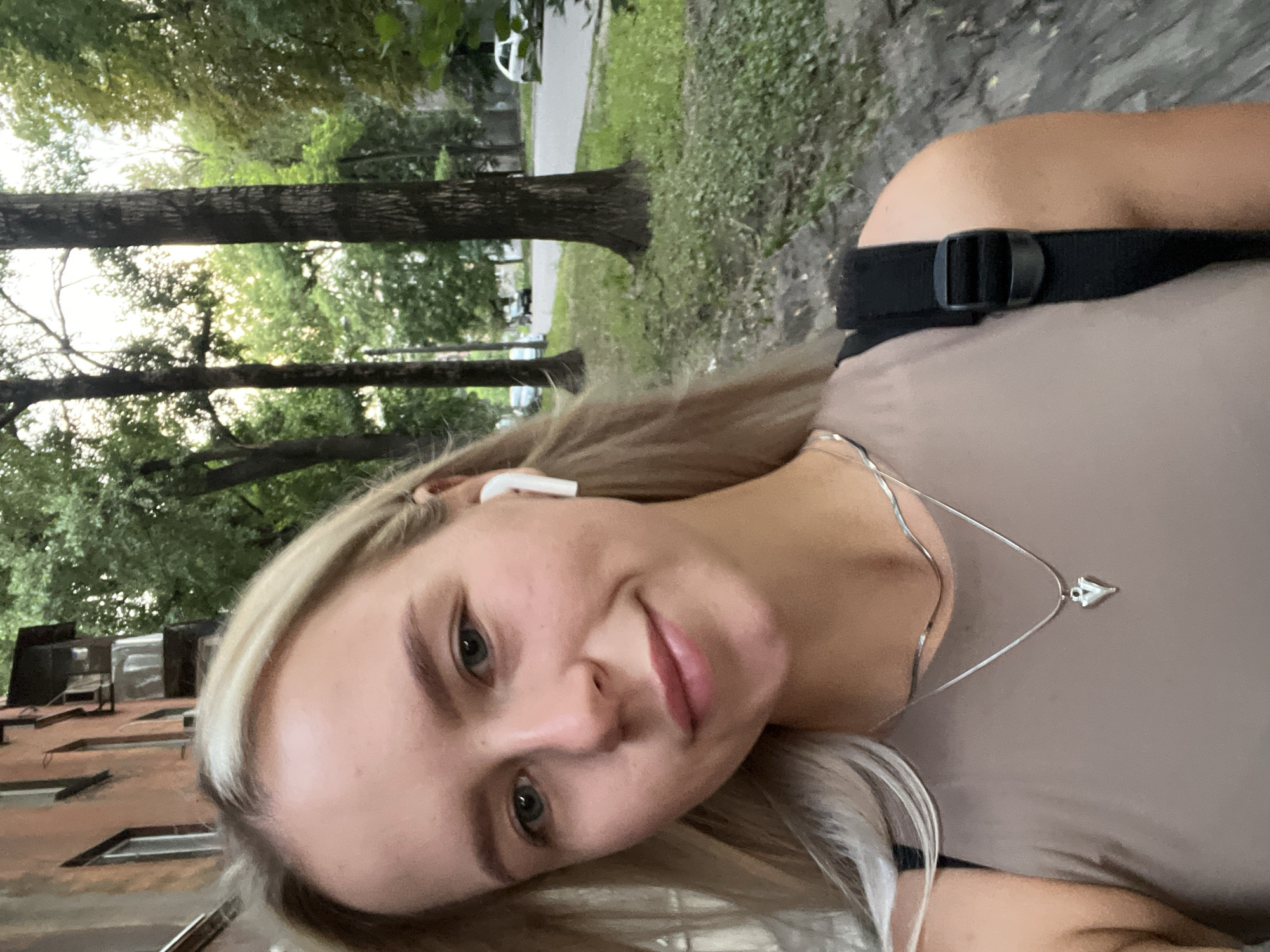 A young blonde woman with blue eyes and fair skin takes a selfie outdoors. She is wearing a tan sleeveless top, a silver necklace with a heart pendant, and white wireless earbuds. A black backpack strap is visible on her right shoulder. She is standing on a sidewalk with a textured gray surface. In the background, there is a multi-story brick building with balconies, tall trees with green foliage, and a street with parked cars. The lighting suggests it is daytime, possibly late afternoon.