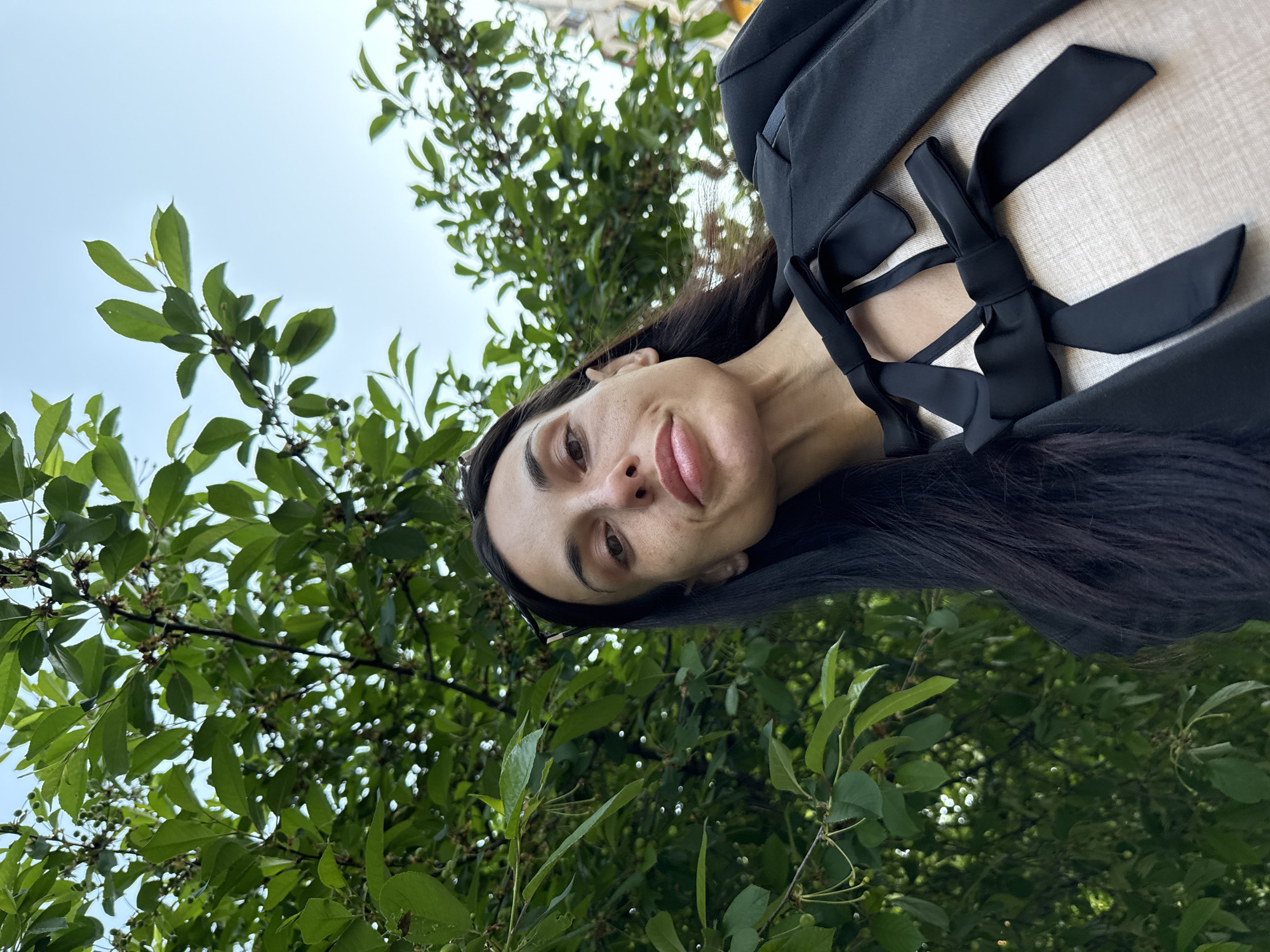 A woman with long, dark hair, wearing a black top with a ribbon design, is viewed from a low angle. She is partially framed by green foliage. The background includes a glimpse of a building and sky. The location cannot be determined from the provided image without further information.