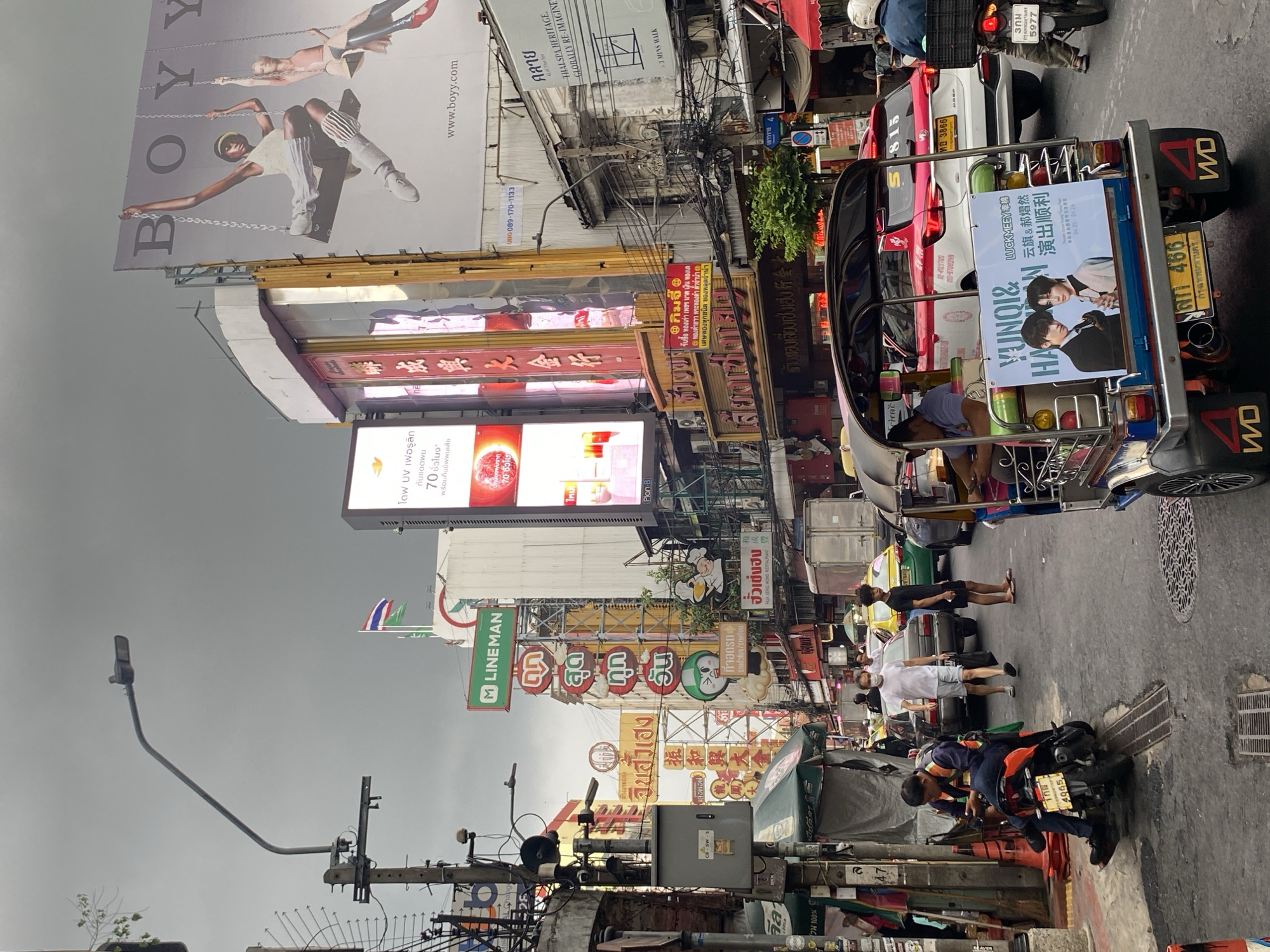 The image captures a street scene in Bangkok, Thailand, characterized by a bustling atmosphere filled with people, vehicles, and a diverse array of signs and billboards.
Several individuals are visible, including pedestrians walking along the street and motorists riding motorcycles. A tuk-tuk is prominently featured.
The urban environment is densely packed with signage in both Thai and English, advertising various businesses, products, and services. Billboards displaying fashion brands and celebrity endorsements are also present.
Electrical wires are tangled overhead, and a street lamp extends out from a utility pole. The overall impression is of a vibrant and chaotic urban landscape in Bangkok.