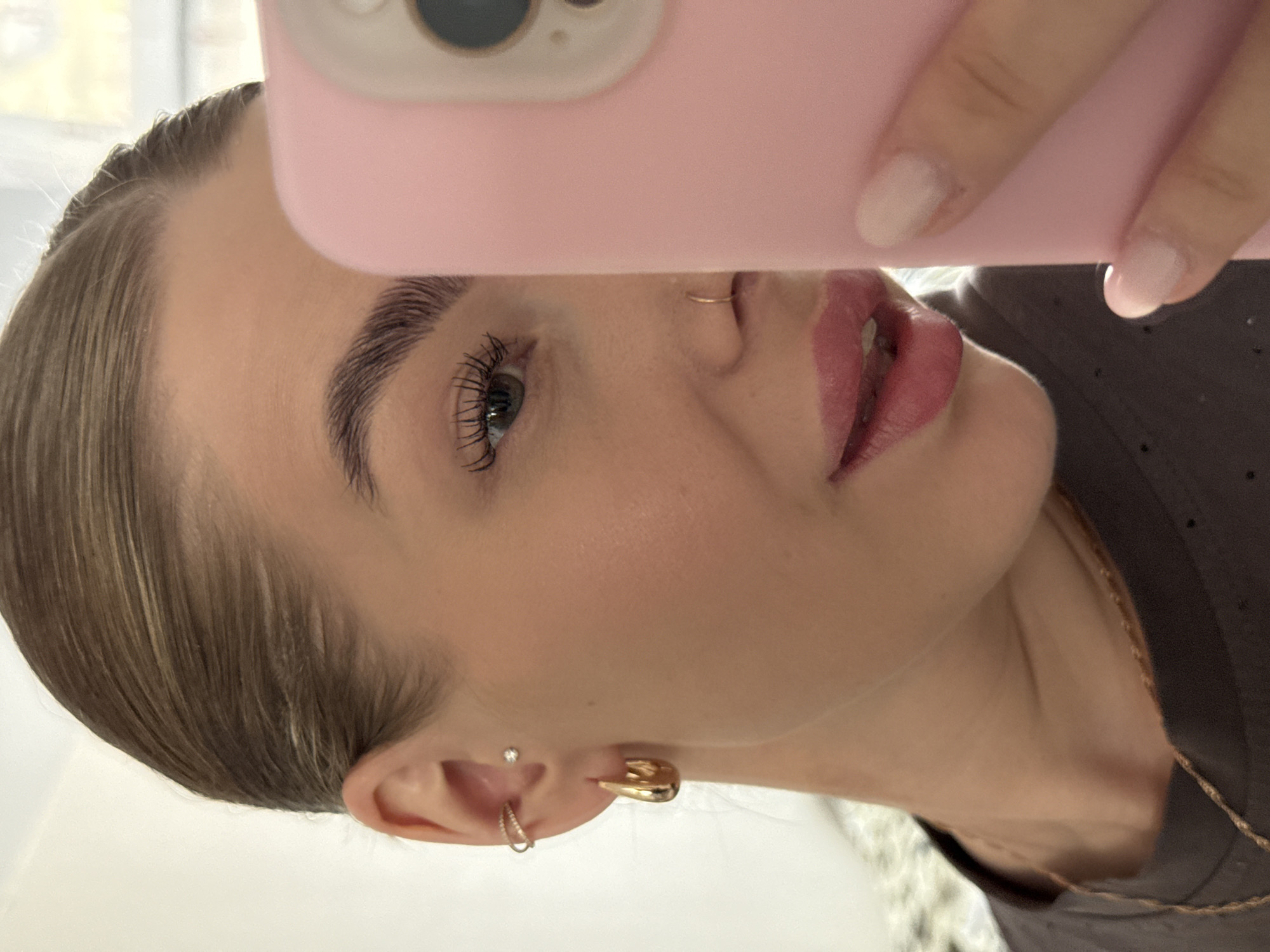 An individual takes a close-up selfie in Chaikovsky, Russia, captured with the image rotated 90 degrees counter-clockwise. The frame focuses on the upper right side of the person's face, partially obscured by a smartphone held by a hand. The phone has a pink case, and parts of its camera lens are visible. The fingernails on the hand are painted with a light, neutral polish.
The person has light-toned skin and light brown hair pulled back. Key facial features include well-groomed, dark eyebrows and a visible blue or grey eye with long, dark eyelashes, enhanced by mascara. A small gold hoop nose ring adorns the nostril. The lips are full, colored with a glossy pink-red lipstick. The visible ear is accessorized with multiple earrings: a small stud, a delicate hoop in the upper lobe, and a prominent gold cuff or hoop. Around the neck, a thin gold chain necklace is visible, paired with a dark-colored top. The background is softly blurred, indicating an indoor setting with subtle natural light, possibly from a window, though specific location cues within Chaikovsky are not discernable. There is no visible text in the image. The overall impression is a deliberate, styled portrait focusing on beauty details and accessories.