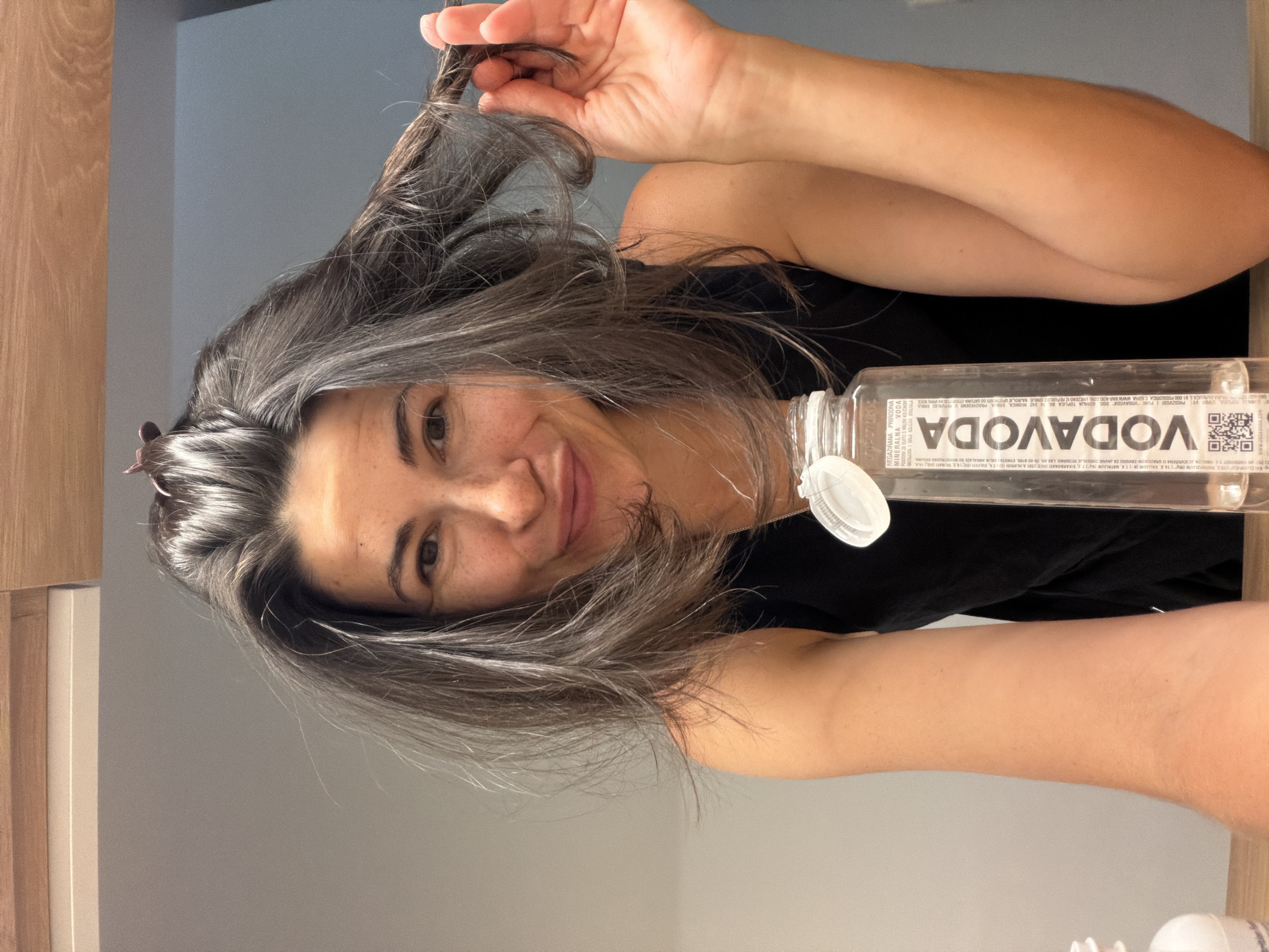 This image is a selfie of a woman taken indoors, likely in Budva, Montenegro. The woman is positioned to the left of the frame, looking directly at the camera with a gentle smile. She has long, dark hair with noticeable gray or silver streaks throughout, and she is holding a section of her hair up with her right hand, while other parts are secured with small clips. Her hair appears somewhat damp or freshly washed and styled. She is wearing a simple black sleeveless top.
To her right, a transparent plastic bottle of "VODAVODA" mineral water is prominently displayed, standing upright. The bottle has a white cap and its label clearly shows the brand name "VODAVODA" printed vertically. The label also features a QR code and text in a Slavic language. The OCR analysis confirms it is "MINERALNA VODA" (mineral water) and "NEGAZIRANA PRIRODNA" (non-carbonated natural), listing components such as potassium, sodium, and bicarbonates. The label also references "GRADSKOM ZAVDOU ZA JAVNO ZORAVLJE REDGRAD LAB," indicating testing by a public health institution in Belgrade.
The background consists of a plain gray wall, with a wooden architectural element visible on the far left, suggesting a clean and modern interior setting, possibly a bathroom or dressing area. The lighting is soft and even, creating a casual and relaxed atmosphere.