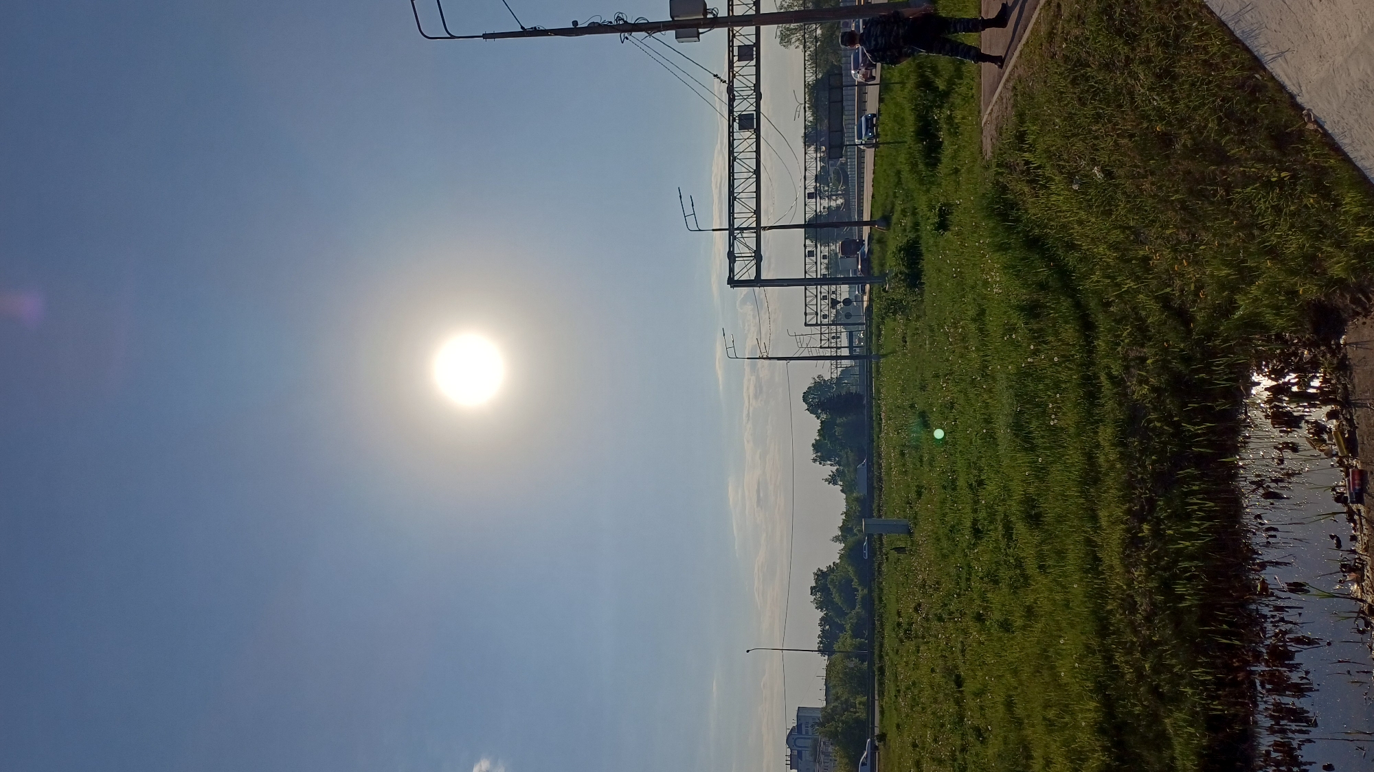 The image depicts a sunny day in Obukhovo, Russia. The sun is high in a clear, light blue sky. A grassy area runs along the right side of the frame, adjacent to what appears to be a road or highway, with occasional standing water reflecting the light near the edge. Utility poles and some kind of steel structure run along the right side of the road, adjacent to the field. A person is walking on the sidewalk along the right edge of the image. Trees and distant buildings are visible on the horizon. Several cars can be seen on the road.