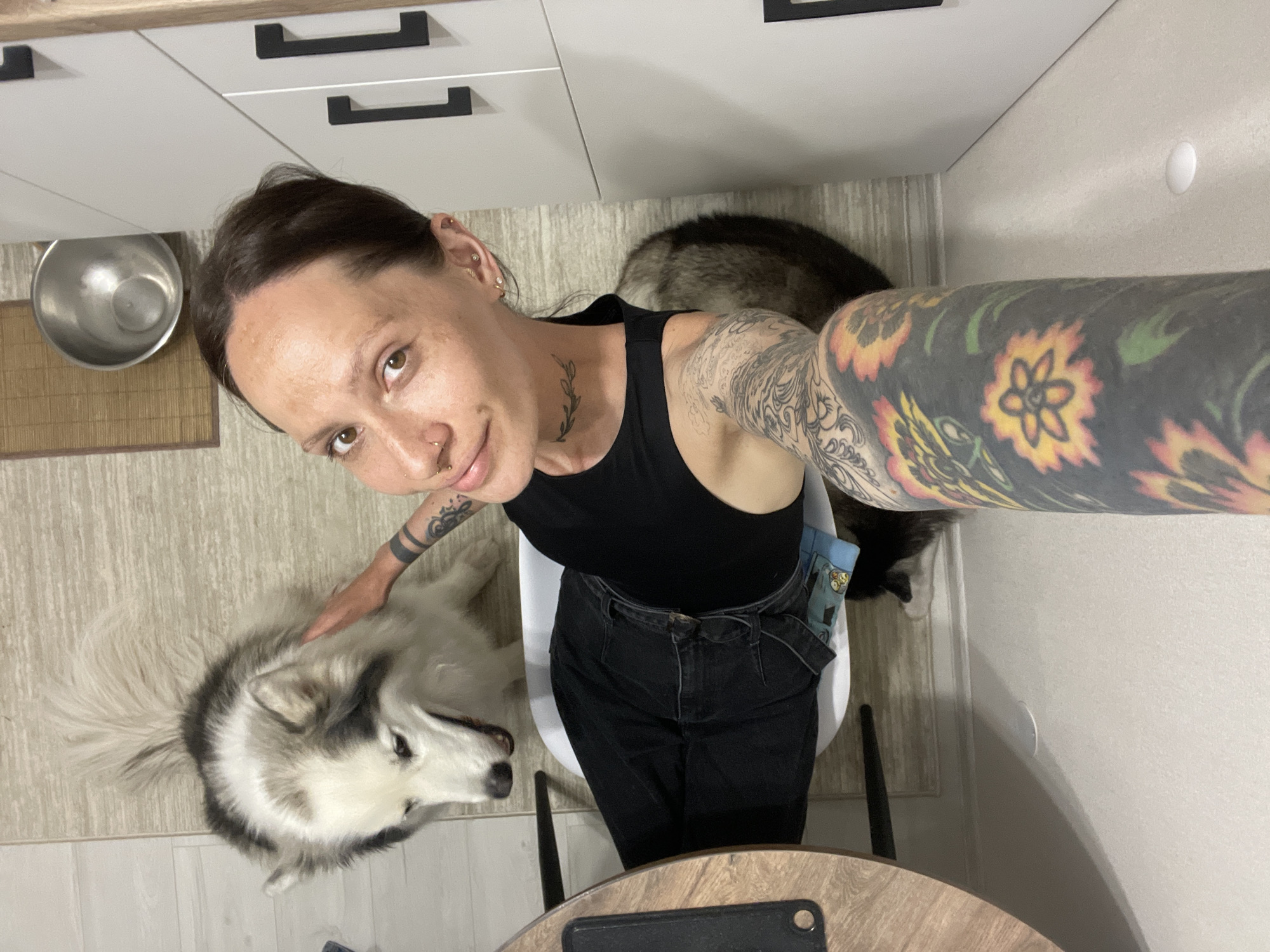 A woman with multiple tattoos and piercings is posing for a selfie indoors with two dogs. She is seated in a white chair and is wearing a black tank top and black jeans. The left sleeve of her arm is heavily tattooed with floral designs. The dogs appear to be huskies. One is lying on the floor near her and is being petted. The second one is behind her. The photo is taken from a high angle, showing a portion of a kitchen with light-colored cabinets with black handles, a stainless steel bowl, and a wooden table. No information can be extracted from the image that confirms that the location is in the city of Izhevsk, Russia.