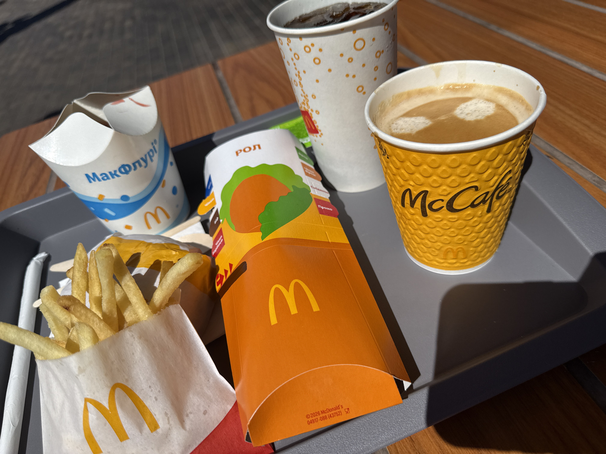 A McDonald's tray sits on a wooden table outdoors. On the tray are a white paper cup with a design and the "McDonald's" logo, a white paper bag with french fries and the "McDonald's" logo, an orange box with the "McDonald's" logo and a graphic of food, a white cup with orange circles and the "McDonald's" logo, and a yellow cup with a textured pattern and the "McCafé" logo. The McCafé cup contains coffee with foam. The white cup contains a dark beverage with ice. The overall scene suggests a meal purchased from McDonald's. The image was taken in Hadzynka, Ukraine.