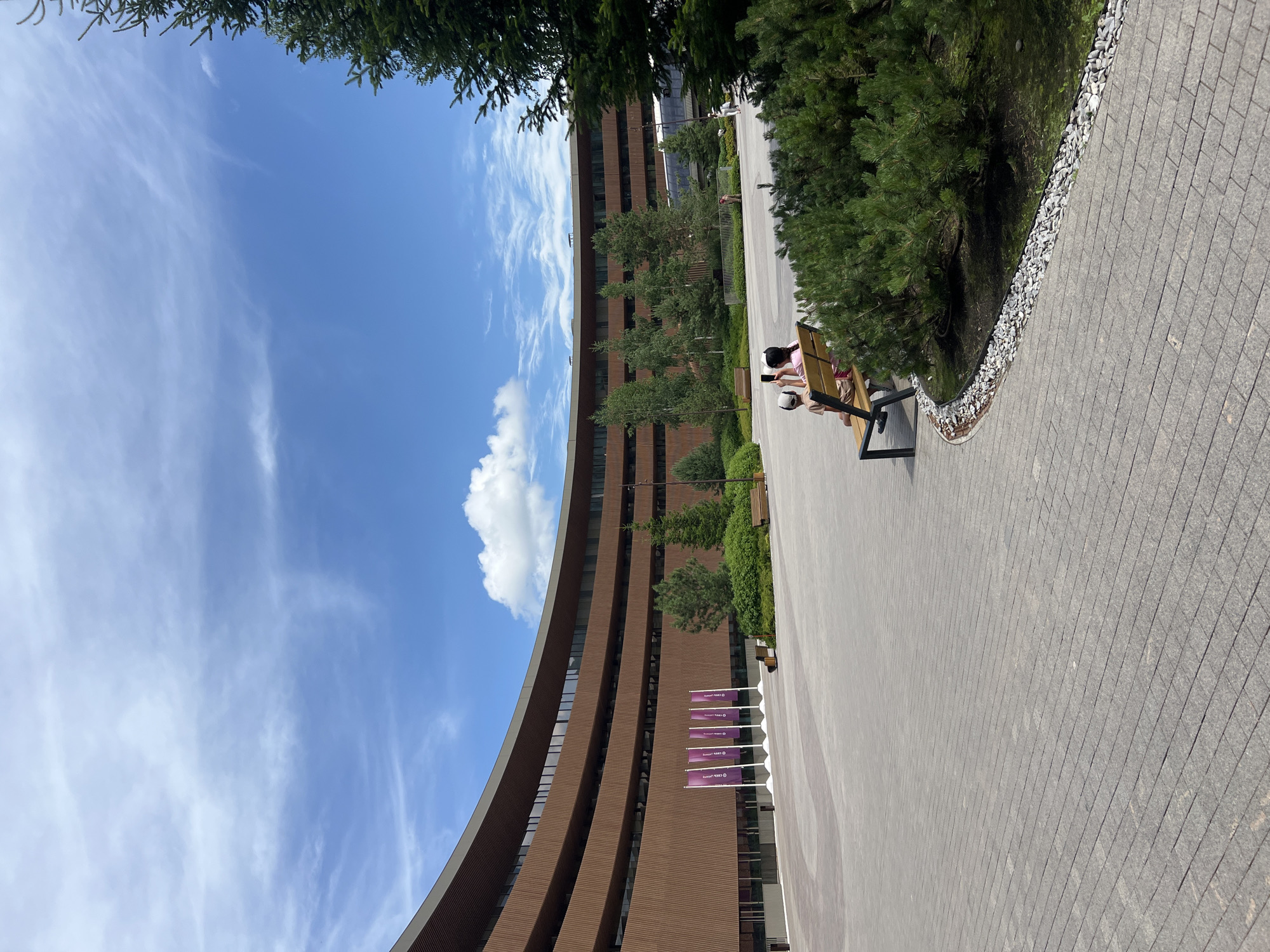 In an open, paved area in front of a curved, multi-story building with a brown, textured facade, two individuals are seated on a wooden bench. The individual on the left wears a light-colored cap and a t-shirt. The individual on the right is partially obscured but appears to be wearing a pink top and is holding a smartphone. They are positioned in the foreground, with a cluster of coniferous shrubs to their right. The building behind them has numerous windows across its upper levels and a series of purple flags are displayed in front of the lower level on the left side of the image. The sky above is blue with scattered wispy white clouds. A large coniferous tree is visible on the far right edge of the frame. The ground is paved with grey rectangular tiles.