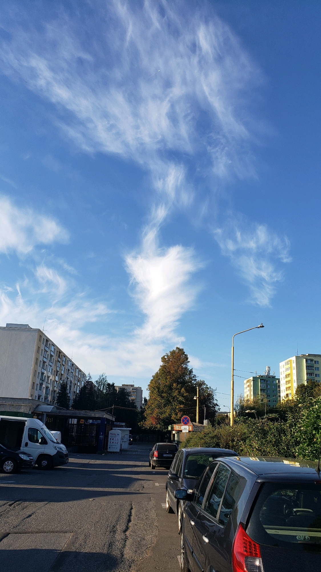 This is an exterior, eye-level, vertical shot of a street scene in Prešov, Slovakia. The sky is a bright blue with wispy white clouds. In the foreground, several cars are parked on asphalt. One car is a dark grey hatchback, facing away from the camera, with its rear bumper and taillight visible. Another dark grey sedan is parked to its left, partially obscuring it. Further back, a black sedan is parked, facing away from the camera. To the left of the parked cars, a white van with its rear door open is parked. Behind the van, there is a row of buildings, including a tall apartment complex with many windows. Further down the street, more buildings are visible, some with light-colored facades and others with colored sections. Trees with green and brown foliage are scattered throughout the scene, particularly to the right of the frame. A street light with a bird perched on top is visible to the right of the frame, along with a traffic sign. The overall impression is a typical urban street scene on a clear day.