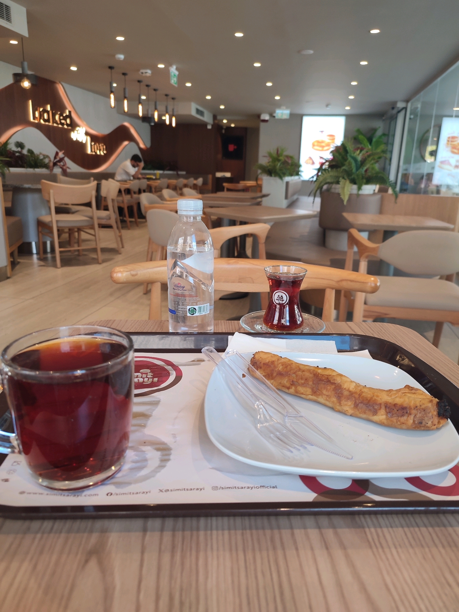 The image shows a tray on a wooden table in what appears to be a cafe or restaurant. On the tray, there are two glasses of tea, one larger glass on the left and a smaller, tulip-shaped glass on the right. Next to the smaller glass is a bottle of water. A single pastry, appearing to be a long, golden-brown roll, is placed on a white plate. A pair of clear plastic forks rests on the plate beside the pastry. The background reveals the interior of the establishment, with wooden chairs and tables, plants, and decorative lighting. A sign with the words "baked with love" is visible on the wall in the background. The overall impression is of a casual dining or cafe setting. The presence of the specific tea glasses, the pastry, and the "simitsarayi.com" URL on the tray liner suggest this location is likely a Simit Sarayı branch, a popular Turkish bakery and cafe chain with numerous locations, including in Kadıköy, Istanbul, Turkey.