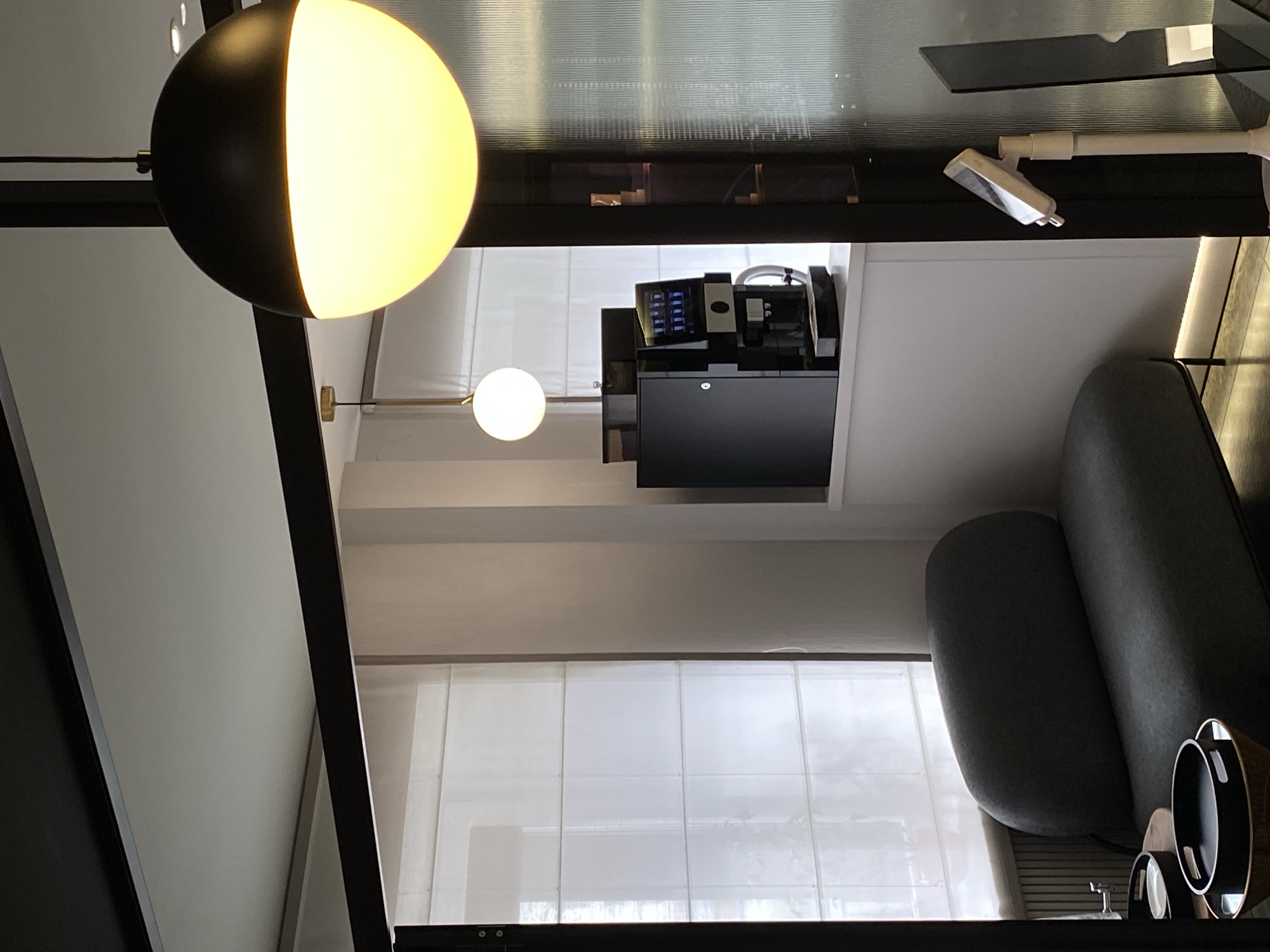 The image shows a room interior with a modern design. Dominating the frame is a spherical pendant lamp with a two-toned color scheme, partially illuminated, adding a contemporary aesthetic. The room appears to be well-lit, possibly by natural light filtering through sheer curtains that cover a window behind a chair or couch. There is a coffee machine situated on a surface. Given the lack of specific indicators, determining the location within the city of Votkinsk, Russia, is impossible based solely on the visual information.