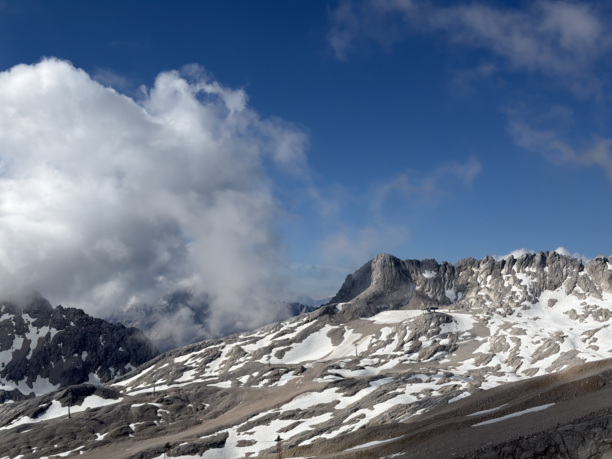 This is a landscape photograph taken from a high vantage point in the Zugspitze massif, the highest peak in Germany, near Garmisch-Partenkirchen. The image depicts a rugged mountain terrain with patches of snow and exposed rock. The sky is a vibrant blue, with dramatic, large white clouds dominating the upper left quadrant of the image. In the mid-ground, a ski lift station with a small building and support structure is visible on a snowy slope. There are also ski lift towers and cables crisscrossing the mountainous landscape. The overall impression is one of a remote, high-altitude environment under a dynamic sky.