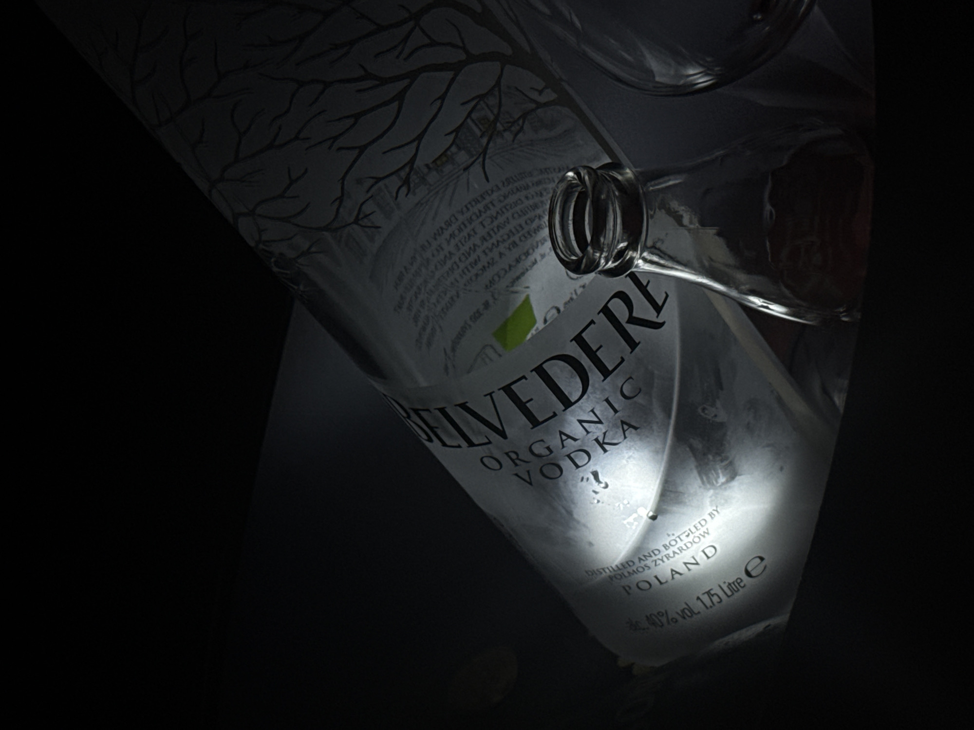 A close-up, low-light photograph depicts an almost empty bottle of Belvedere Organic Vodka. The bottle is angled diagonally across the frame, with its neck toward the upper-right corner. The label is partially illuminated, revealing the brand name "BELVEDERE ORGANIC VODKA" in large letters, along with details such as "DISTILLED AND BOTTLED BY POLMOS ZYRARDÓW POLAND" and the alcohol content "40% vol. 1.75 Litre e". A tree branch design is visible on the upper portion of the bottle. The background is dark and indistinct, with only the bottle and its immediate surroundings visible. No information on the location in the city of Sofia, Bulgaria, is available in this image.