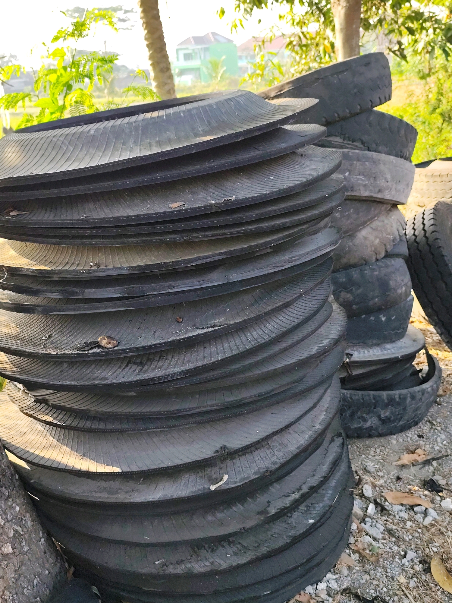 A stack of black, worn tires is piled outdoors in Jebugan, Indonesia, with a building visible in the background. The tires are layered on top of each other, with some appearing flatter than others. Sunlight filters through trees, casting shadows on the ground. The ground beneath the tires is covered with gravel and dry leaves.