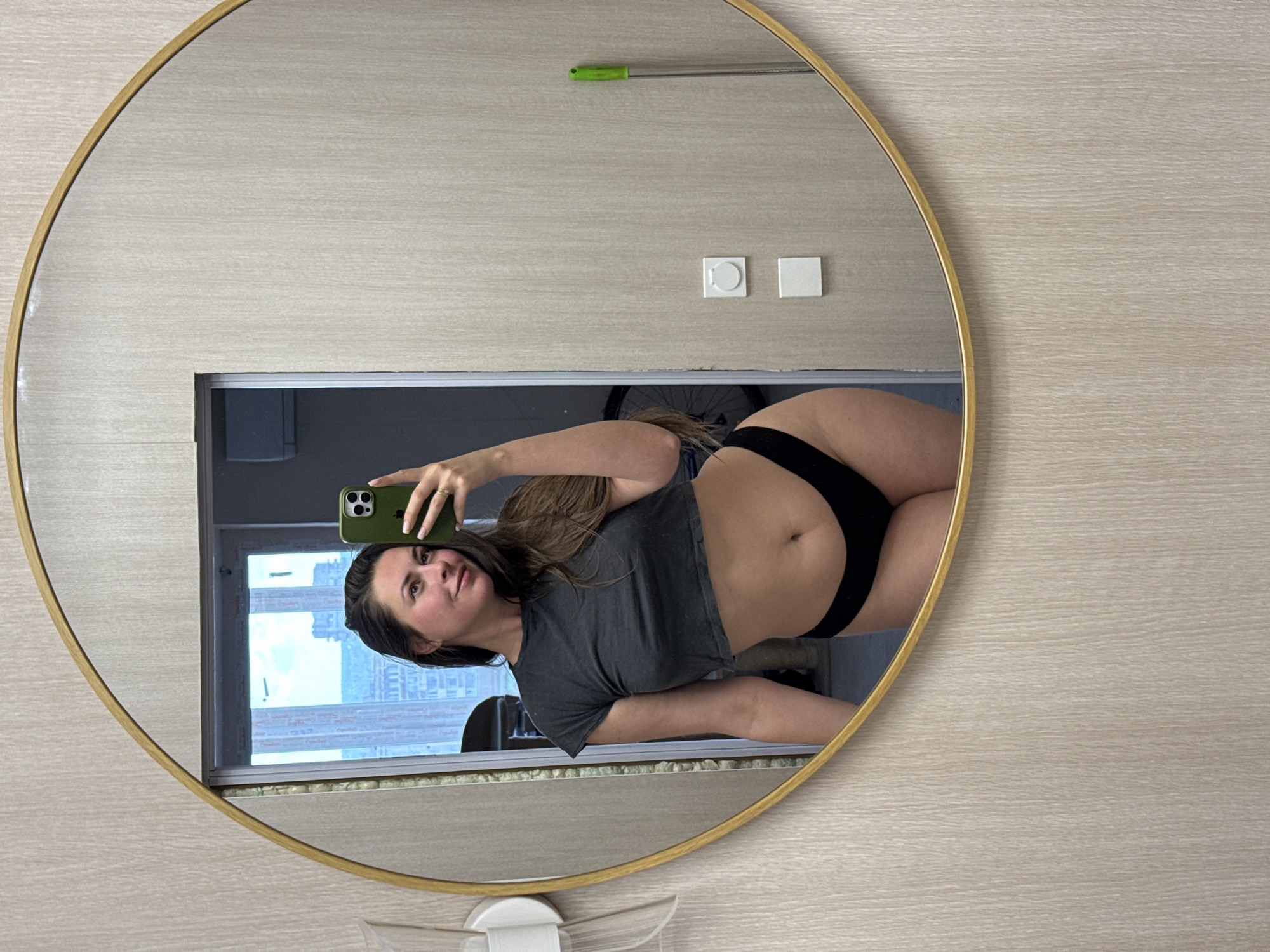 A woman with long brown hair takes a selfie in a round mirror. She is wearing a dark gray cropped t-shirt and black underwear. The mirror is framed with gold and is hanging on a wall with a light wood grain pattern. In the reflection, a window is visible, showing a cityscape with tall buildings. Other objects visible in the reflection include a white light fixture on the left, a bicycle wheel, a green-handled mop on the right, and white electrical outlets on the wall. The woman is smiling and looking at her phone, which is green and held in her right hand.