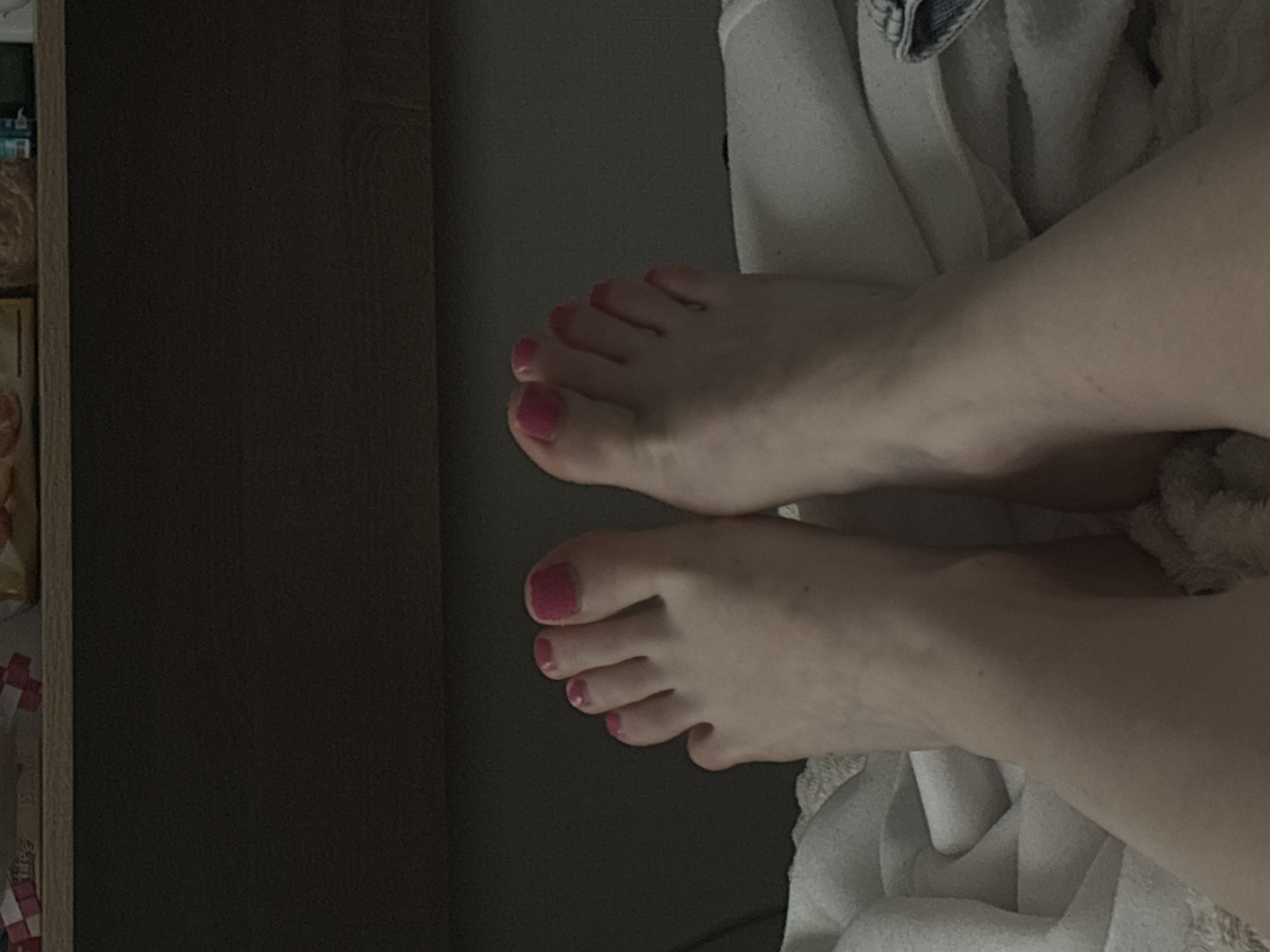 The image shows two Caucasian female feet with pink nail polish. The feet are resting on a white towel. The background includes a dark gray wall and a partial view of a wooden cabinet. The location cannot be determined to be Vsevolozhsk, Russia based on the image alone.