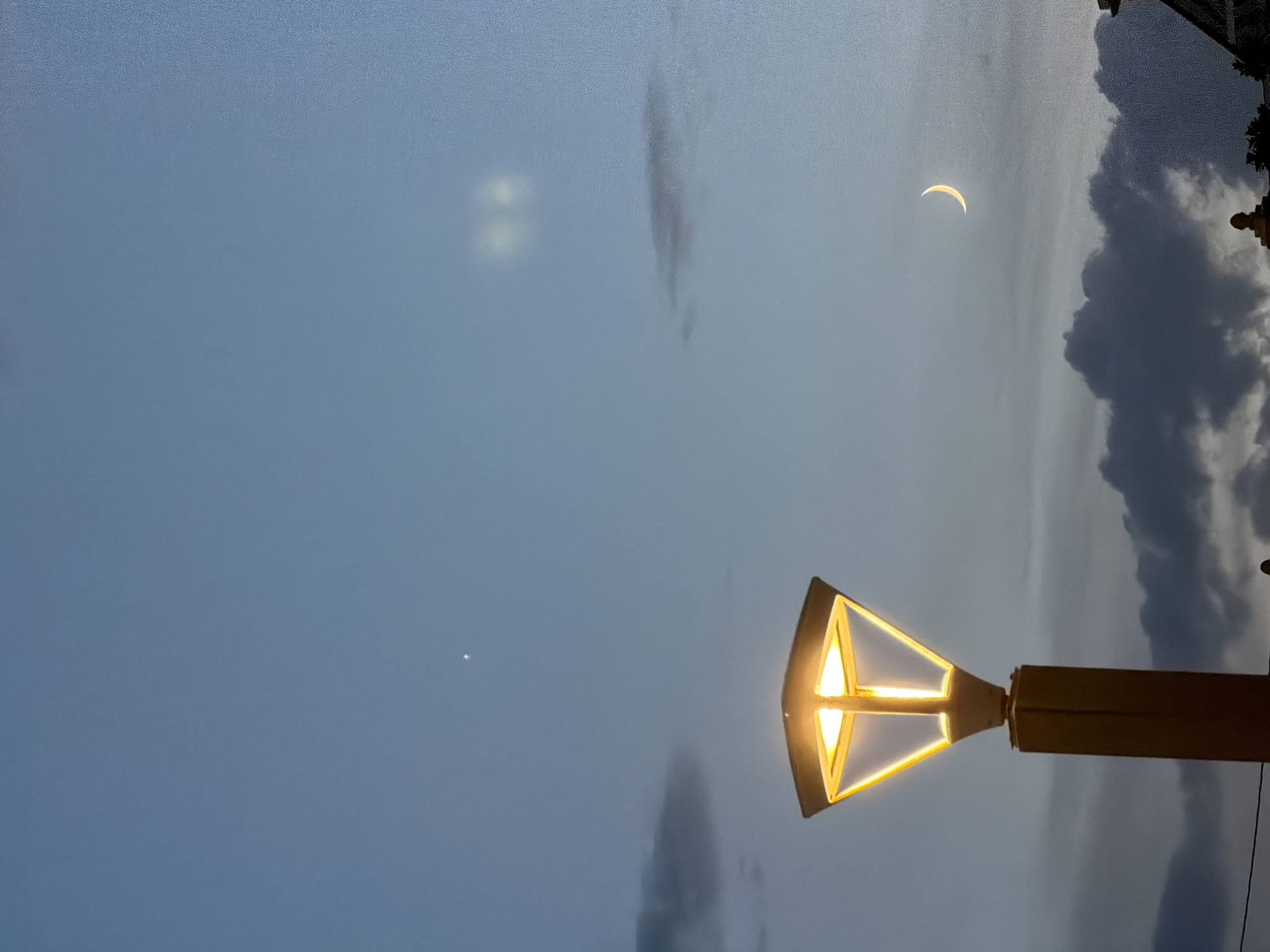 The image captures a twilight scene in El Campello, Spain, featuring a contemporary streetlamp with a yellow, angular luminaire emitting a warm glow. The sky is a dusky blue, with scattered clouds ranging from dark, heavy formations near the horizon to lighter, wispy ones higher up. A thin crescent moon is visible in the upper right quadrant of the sky, positioned above a dark, elongated cloud. Slightly to the left of the moon, a bright, star-like object is present, possibly Venus. In the far upper left corner, another faint, star-like object is visible. The streetlamp stands prominently in the foreground on the left side of the frame, with its yellow post extending downwards. The base of buildings or structures can be seen at the very bottom of the image, indicating an urban or semi-urban environment.