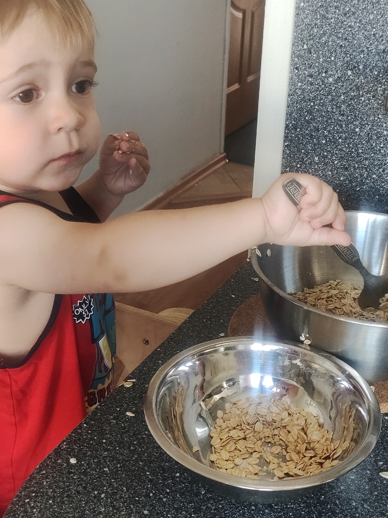 A young child with blonde hair and brown eyes is shown in a close-up shot in a kitchen. The child is wearing a red tank top with a graphic design. The child's left arm is extended, holding a spoon that is in a large metal bowl filled with oatmeal. Another smaller metal bowl with oatmeal is in the foreground. The kitchen counter is dark and speckled. The background shows a wall and a doorway. There are no explicit indicators of the specific location within Chaikovsky, Russia.