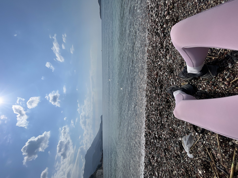 A first-person perspective photograph depicts the lower body of an individual seated on a pebble beach in Rafailovići, Montenegro, facing the sea. The subject's legs are extended, clothed in light purple leggings, black athletic shoes, and white socks. The immediate foreground comprises diverse dark and light pebbles, interspersed with dry plant matter and a crumpled white plastic bag near the left leg. The clear, calm sea in the midground displays a turquoise tint with minor wave activity at the shoreline. The background on the left reveals a mountainous coastline dotted with several multi-story buildings, while the right features open sea with a small, dark vessel on the distant horizon and a faint landmass to the far right. The sky is bright blue with scattered white cumulus clouds, and the sun, positioned in the upper left, creates a prominent lens flare.