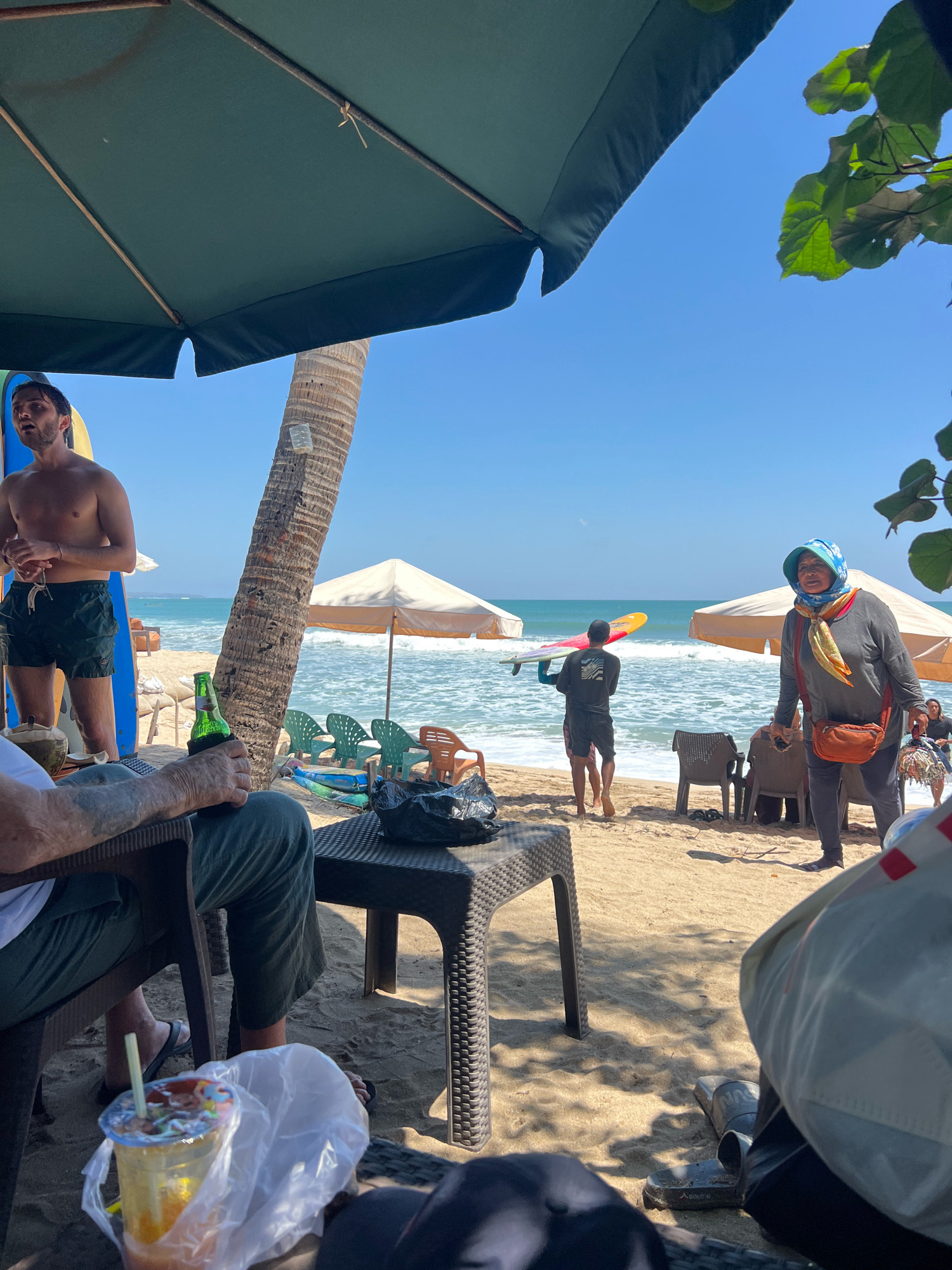 A group of people are relaxing on a beach in Kuta, Indonesia, under a large green umbrella. In the foreground, a man with tattoos on his arm holds a bottle of beer while sitting in a chair. Next to him is a table with a drink and a plastic bag. To the left, a shirtless man with dark hair stands near a surfboard. In the background, a person carrying a surfboard walks towards the ocean, where waves are breaking. A woman wearing a headscarf and a red purse walks on the sand, and several beach chairs and umbrellas are visible. The sky is clear and blue, and the sun is shining.