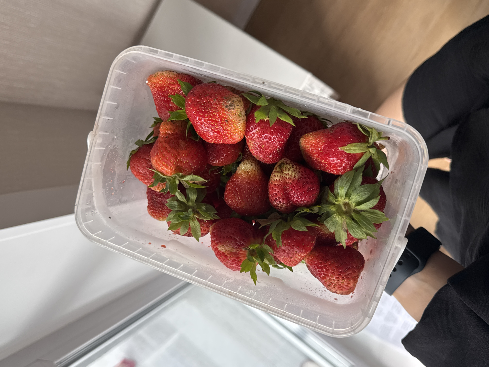 A person is holding a clear plastic container filled with ripe strawberries. The container is tilted, with the strawberries spilling slightly over the edge. The background is a blurred indoor scene, suggesting a home environment. The lighting appears to be natural, coming from a window to the left. The person is wearing black clothing and a dark smartwatch on their left wrist. There is no indication of the specific location within Perm, Russia.