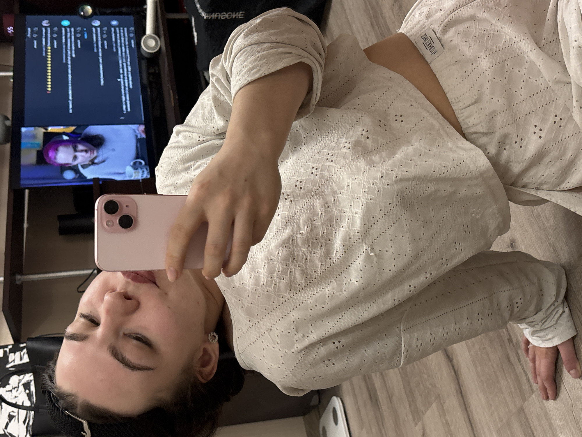 A person with dark hair is lying on a wooden floor, taking a selfie with a pink phone. They are wearing a white, eyelet-patterned top. In the background, there is a television displaying a video call. A woman with purple hair is visible on the screen. A black headset rests nearby, and a black bag hangs in the periphery. A scale is partly visible on the floor near the person's feet. This image was possibly taken in the city of Perm, Russia.