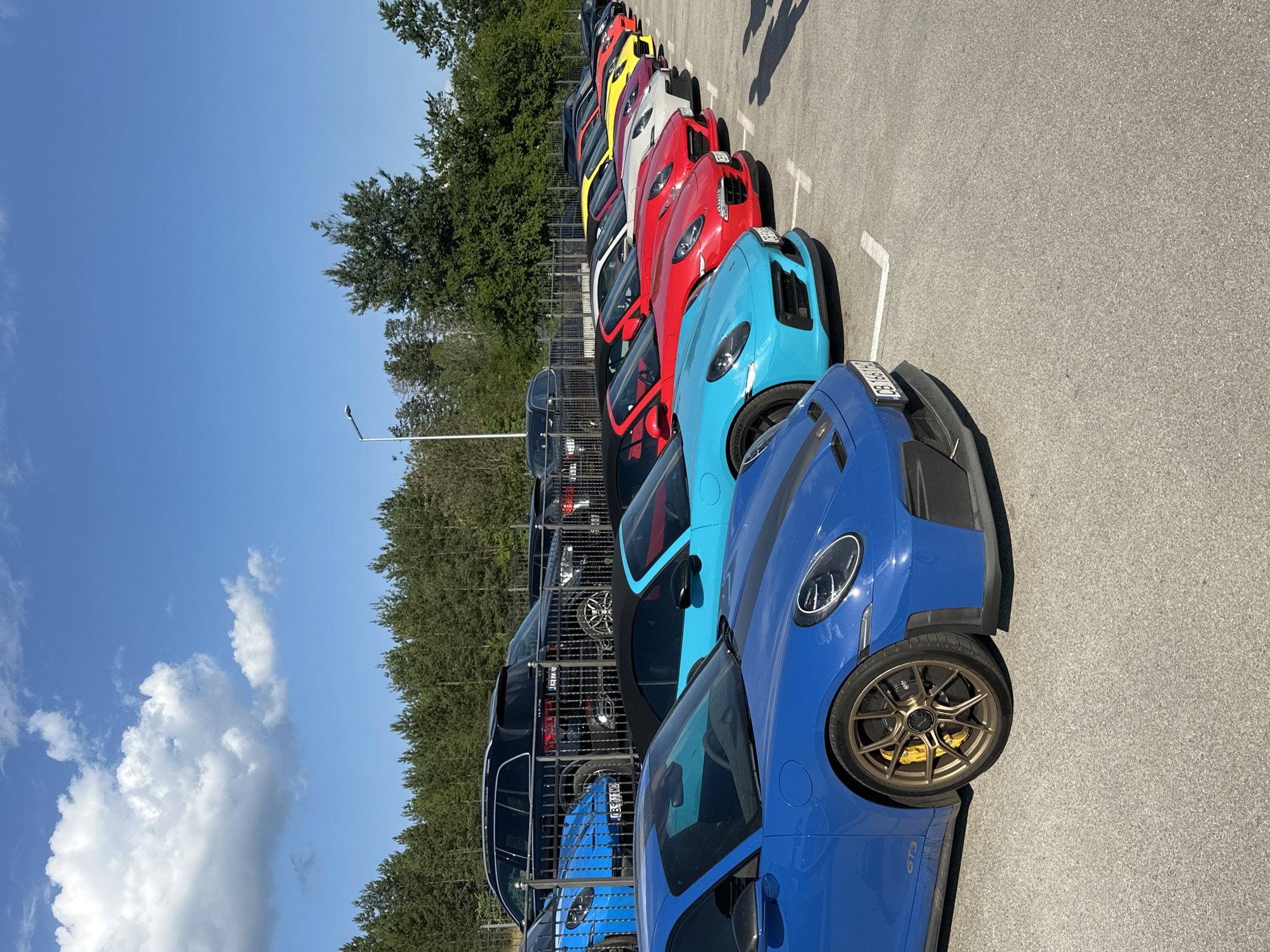 The image depicts a row of colorful Porsche sports cars parked in an outdoor lot. The cars are lined up in a diagonal fashion, with the closest car being a bright blue Porsche GT3. Behind it, several other Porsches are visible in various colors, including aqua, red, white, and yellow. The cars are parked on a paved surface marked with white parking lines. In the background, a dense line of green trees forms a natural backdrop, with a clear blue sky overhead. The scene appears to be an organized car gathering or event. The location is Kerenite, Bulgaria.