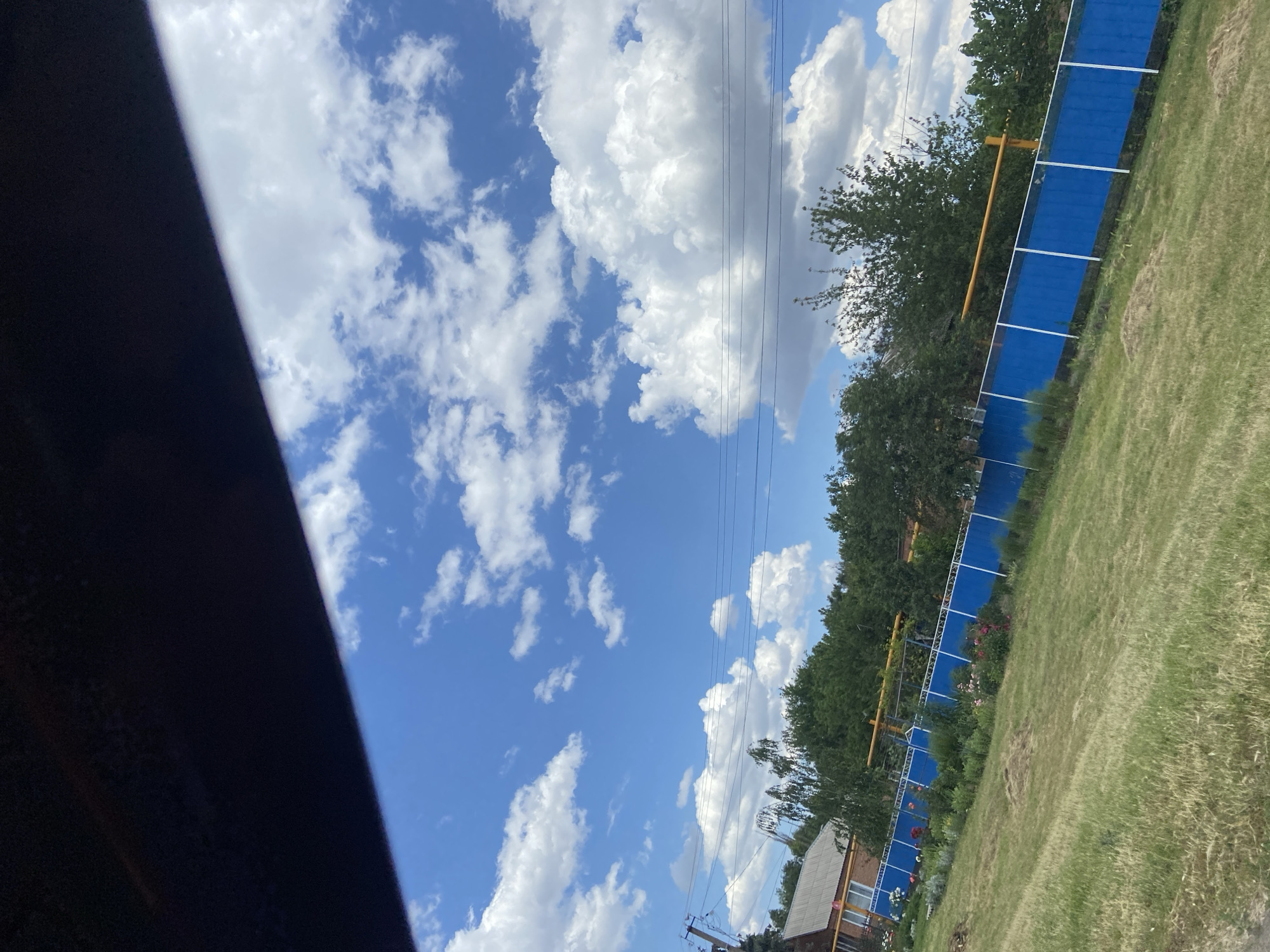 This is a landscape image taken outdoors, with the camera oriented vertically. The sky dominates the upper half of the frame, featuring a clear blue expanse punctuated by fluffy white cumulus clouds. Power lines traverse the sky diagonally from the upper left to the lower right.
The lower half of the image depicts a grassy area with a prominent blue fence running parallel to the right edge. Behind the fence, a line of trees forms a dense green border. A small section of a brick building with a light-colored roof is visible in the lower right corner, near the fence. A yellow structure, possibly part of a fence or pole, is also visible behind the blue fence. The ground in front of the fence is covered in grass, some of which appears dry or sparse. Flowers are visible in a garden bed along the fence line.
The lighting suggests it is daytime, likely a sunny day given the bright sky and well-defined clouds. The scene appears to be a rural or suburban setting, possibly a backyard or park area, in Tagarog, Russia. There is no discernible activity or event taking place, and no visible text. The image evokes a sense of calm and openness due to the expansive sky and greenery.