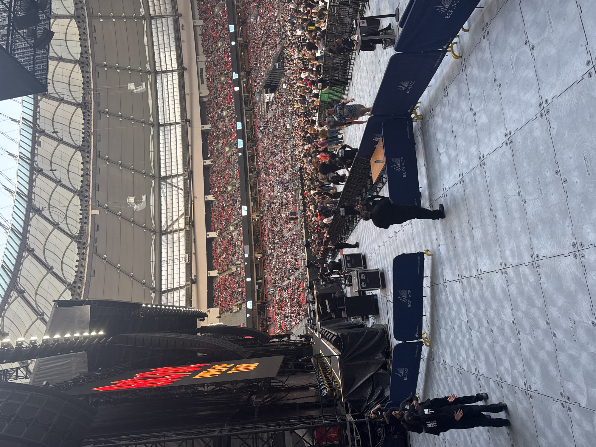 This image depicts a large concert or sporting event taking place inside BC Place Stadium in Vancouver, Canada. The stadium's retractable roof is partially open, allowing natural light to filter into the arena. A massive stage, adorned with large black speakers and a bright red digital display that reads "WORLD TOUR," is visible on the left side of the frame.
In the foreground, a grey, tiled floor is lined with dark blue barriers bearing the "BC PLACE" logo. Several individuals, some appearing to be security personnel in black uniforms, are positioned near these barriers and on the stage area. One person in the foreground, wearing a turban and a black shirt with "BC PLACE SECURITY" printed on it, is facing away from the camera.
The stands are filled with a diverse crowd, predominantly wearing red tops, suggesting a common theme or team affiliation. The sheer number of attendees signifies a major event. The overall atmosphere appears to be one of anticipation before a performance or during a break in the action. The environment is clearly an indoor sports and entertainment venue, with a capacity for tens of thousands of spectators.