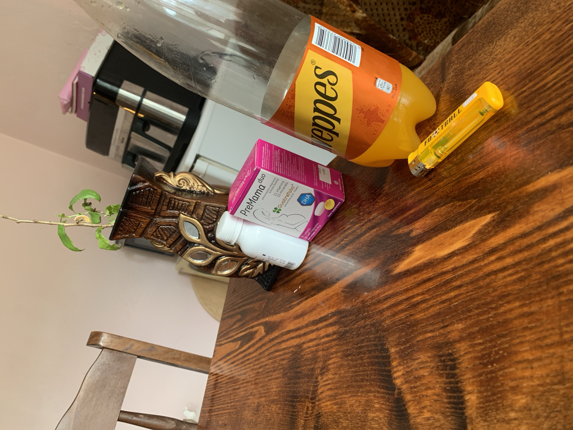 The image depicts a close-up, angled view of a wooden table. On the table, a nearly empty plastic bottle of Schweppes orange soda is prominent. To the left of the bottle, there is a pink and white box labeled "PreMama duo" with some text in Bulgarian, and a white bottle with a white cap. A yellow lighter with "FOOTBALL" printed on it lies in the foreground. In the background, a portion of a wooden chair and a decorative wooden vase with some plant sprigs are visible. A coffee maker and stacked boxes are also partially seen in the background. The lighting suggests an indoor setting, possibly a home. There are no people or explicit actions captured in the image. The location of Krushev Dol, Bulgaria is not visually represented in the image.