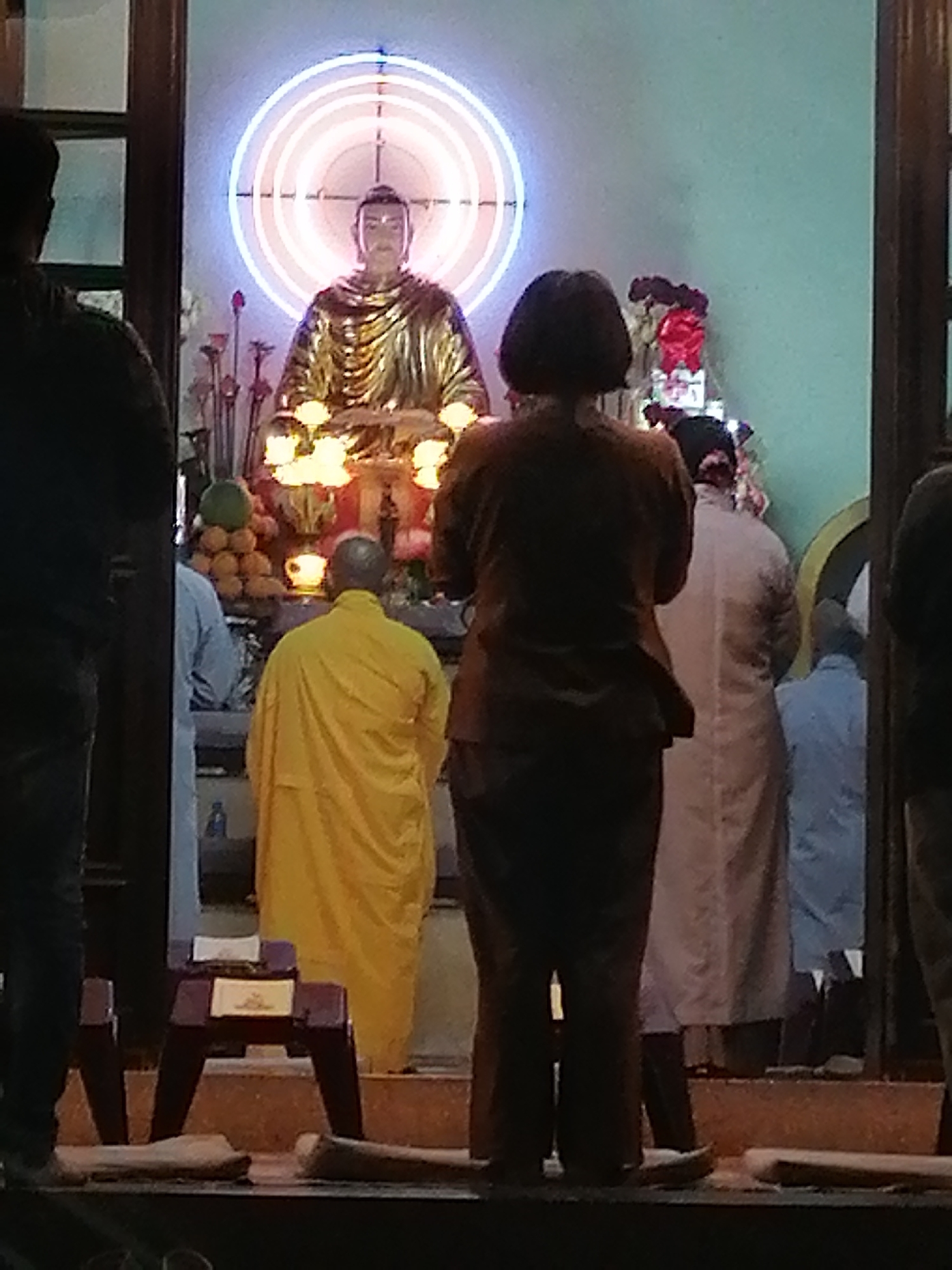 The image depicts the interior of a Buddhist temple in Da Lat, Vietnam, likely during a prayer or ceremony. A golden statue of Buddha, adorned with a glowing circular halo, is the central focus. In front of the statue, several individuals are seen with their backs to the camera, participating in the event. One person is wearing a bright yellow robe, indicative of a monk. Other individuals are dressed in casual attire, with some wearing what appear to be light-colored robes or long garments. The temple's architecture suggests traditional Vietnamese Buddhist design, with wooden elements and possibly a serene, spiritual atmosphere. The lighting is dim, with the Buddha statue and its halo being the primary sources of illumination, creating a contemplative mood. The scene captures a moment of devotion and community within a religious setting.