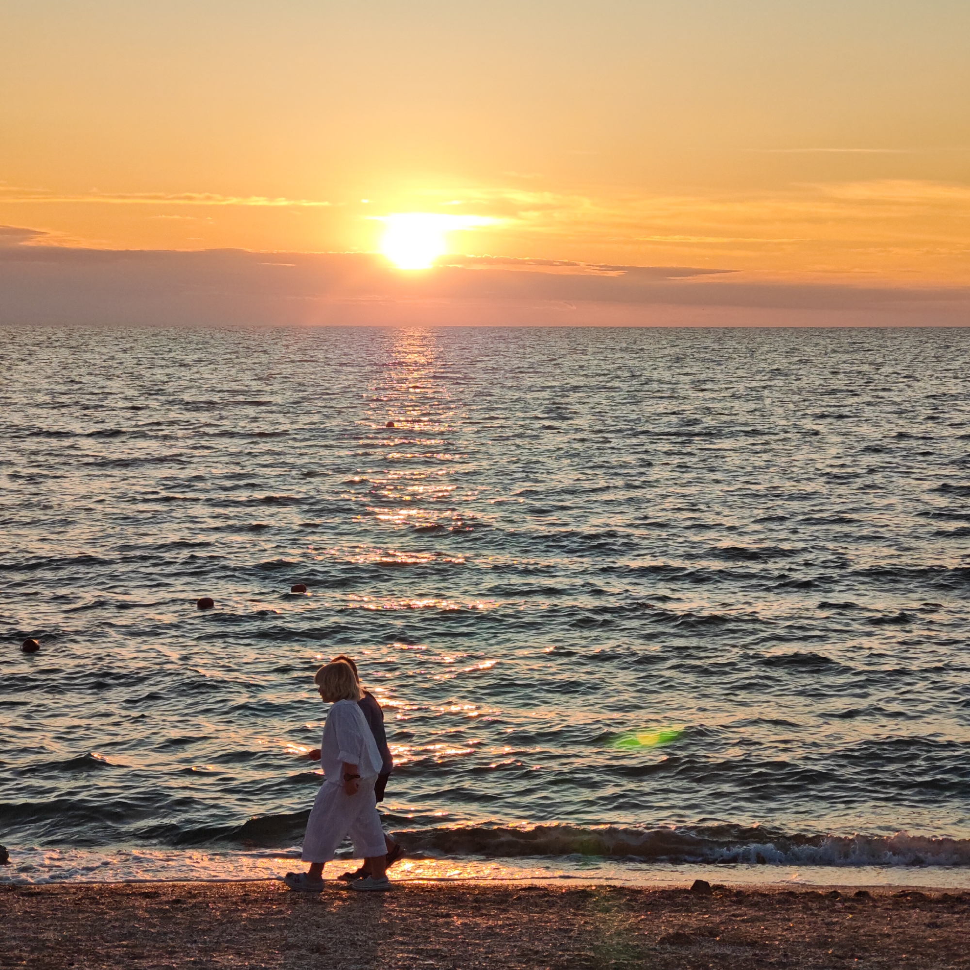 This is a horizontal, full-shot photograph of a sunset over the ocean. The sun is low in the sky, casting a warm, orange glow that reflects off the water. The sky is a gradient of yellow and orange, with some clouds present. Two people are walking along the beach in the foreground, their silhouettes visible against the bright ocean. The beach is made of small pebbles and sand. The water is calm with gentle waves lapping at the shore. There are a few buoys floating in the water. The overall mood of the image is peaceful and serene.