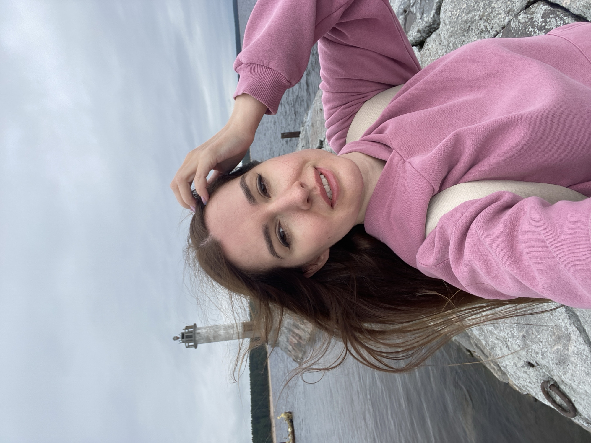 A young woman with long, brown hair, wearing a pink hooded sweatshirt and a backpack, is captured in a selfie. She is standing on a rocky pier next to a body of water. In the background, a lighthouse stands on the opposite shore, with a forest line behind it. The sky is overcast. The location is identified as Моторное, Russia.