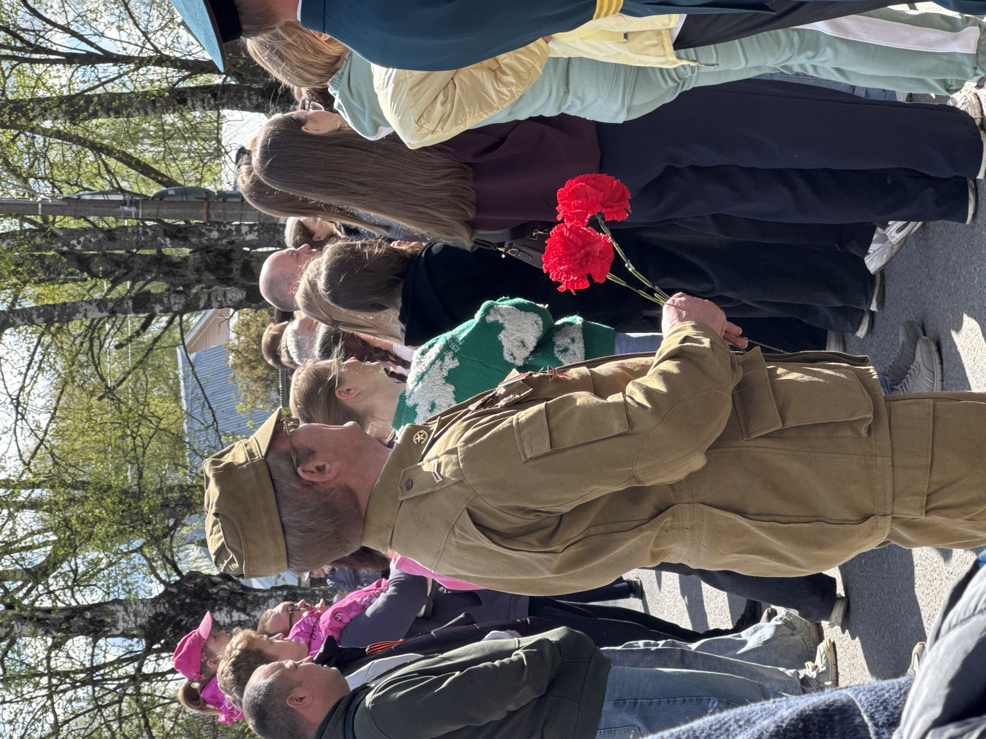 The image depicts a group of people standing together in what appears to be a public space, possibly in the city of Yamyshevo, Russia. The focus is on an elderly man wearing a military-style uniform with decorations and holding two red carnations. Other individuals are seen lined up behind him, suggesting a solemn event or ceremony. The environment shows trees and a building in the background, indicating an outdoor setting during daylight. The people are looking in the same direction.
