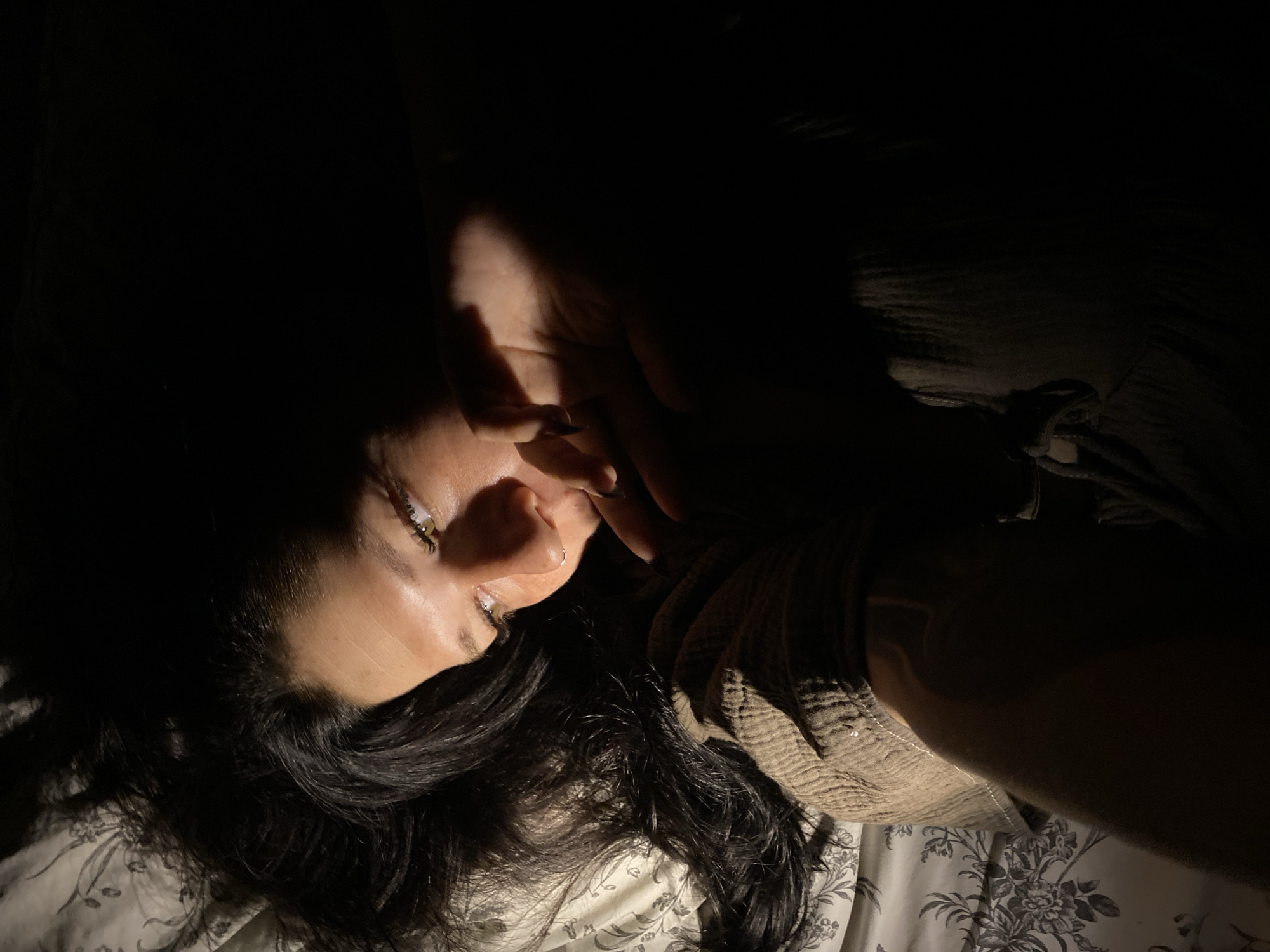 The image depicts a close-up, high-angle shot of a person lying down in what appears to be a bed. The lighting is dim, with a strong light source coming from the upper right, casting dramatic shadows. The person has dark hair and their face is partially obscured by their hand, which is raised near their mouth. Their eyes are open and looking upwards. They are wearing a textured, light brown garment. The background consists of a patterned bedsheet, with a floral or leaf-like design in muted tones. The scene is indoors, likely a bedroom. No specific location in Chaikovsky, Russia is identifiable from the image content.