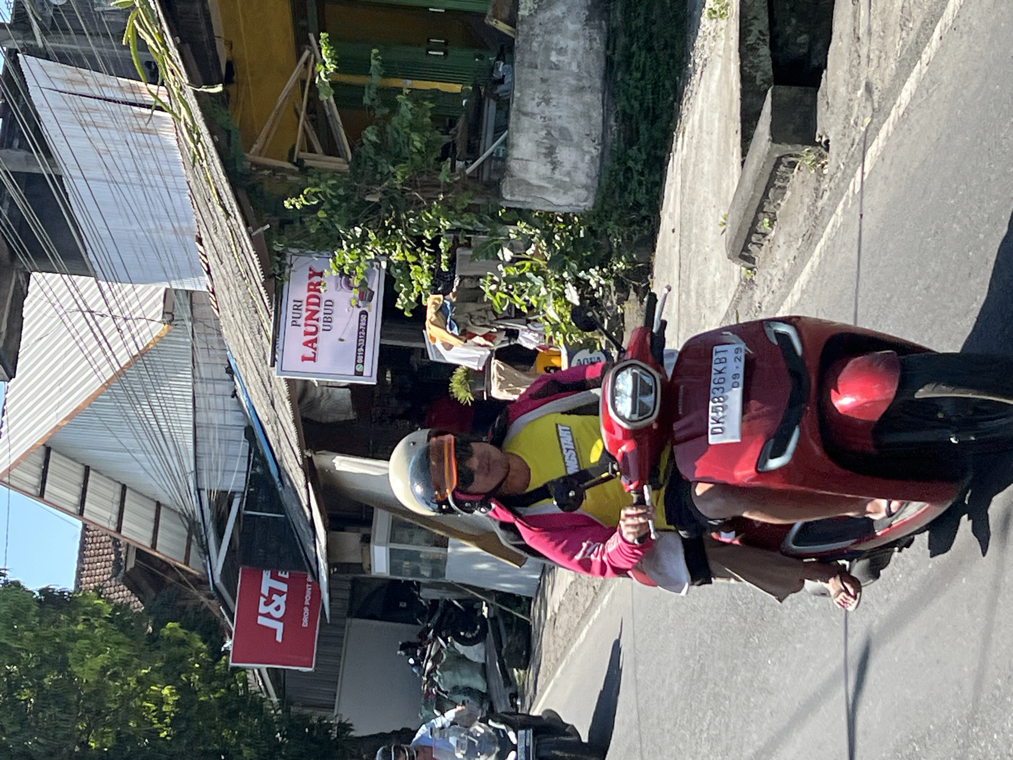 In Ubud District, Indonesia, a person wearing a pink jacket over a yellow t-shirt and sunglasses rides a red scooter. They are wearing a helmet with an orange visor and a black strap. Their legs are visible, with one foot in a sandal and the other in a flip-flop. The scooter has a license plate that reads "DK5836KBT 09-29". Behind the rider, a passenger's legs are visible, also wearing sandals and a brown garment. The scooter is on a paved road, and to the right, a concrete structure with vegetation grows from it. In the background, buildings with corrugated metal roofs are visible, along with signs for "J&T DROP POINT" and "PURI LAUNDRY UBUD". Trees and electrical wires are also present. Another scooter is visible in the left background, ridden by a person wearing a helmet.