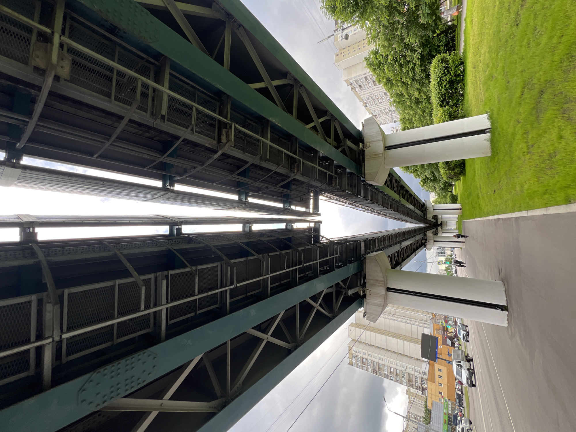 The image depicts the underside of a monorail bridge in Moscow, Russia. The bridge structure is constructed of steel beams and lattice work, painted green. It is supported by white, cylindrical concrete pillars. Above the bridge, there is a glimpse of sky and buildings. Adjacent to the bridge, there's a green grassy area with trees and a sidewalk. Below the bridge, there's a road with cars parked and moving. A pedestrian can be seen walking on the sidewalk near the bridge. The buildings are multi-story residential apartments. The image is taken from a low angle, looking upwards along the length of the monorail bridge.
