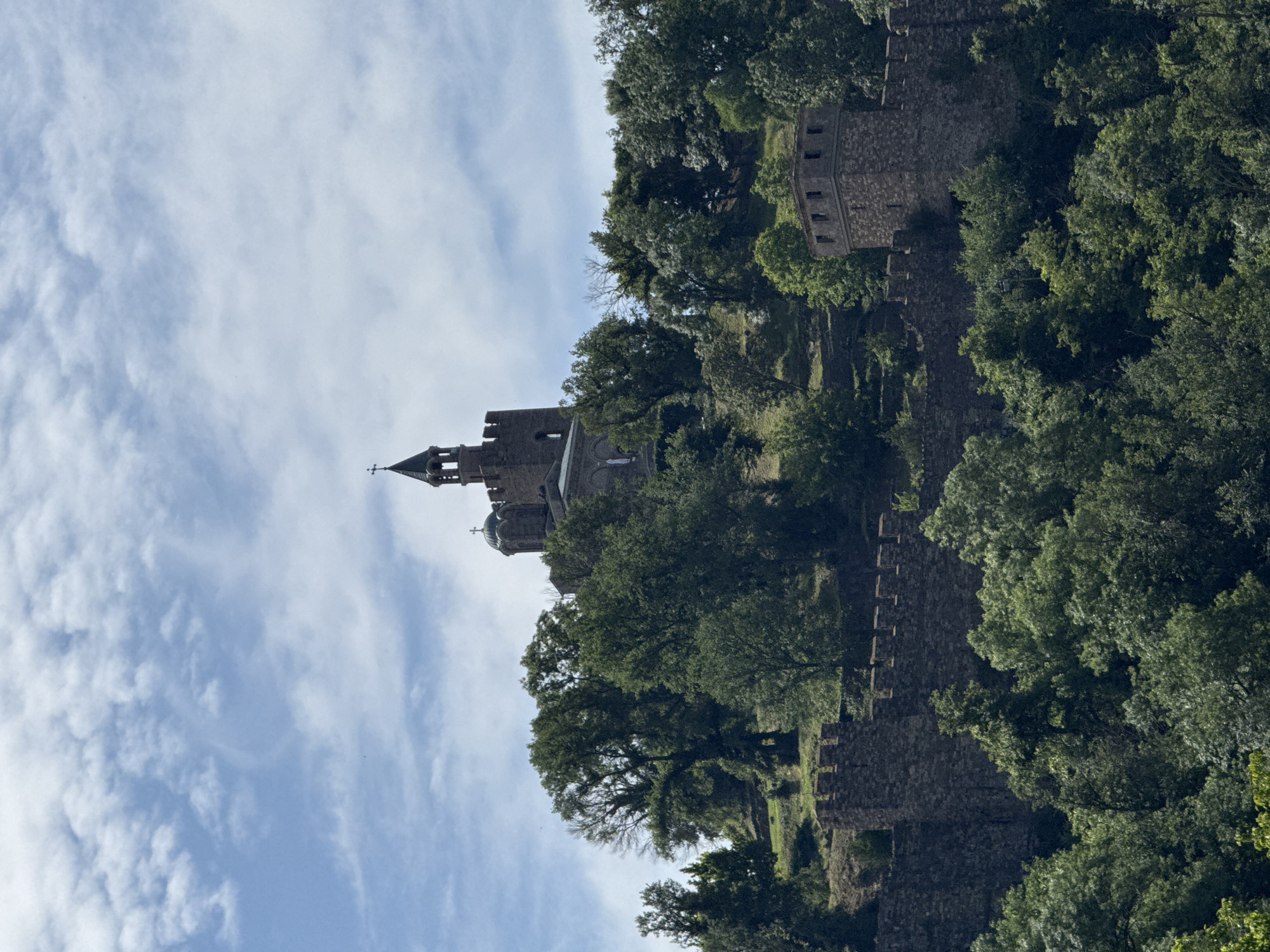 The image shows the Tsarevets Fortress in Veliko Tarnovo, Bulgaria, viewed from a low angle. The fortress walls, made of stone, are visible, partially obscured by trees. The Patriarchal Cathedral of the Holy Ascension of God stands prominently within the fortress, its tower reaching towards the sky. The sky is light blue with wispy white clouds.