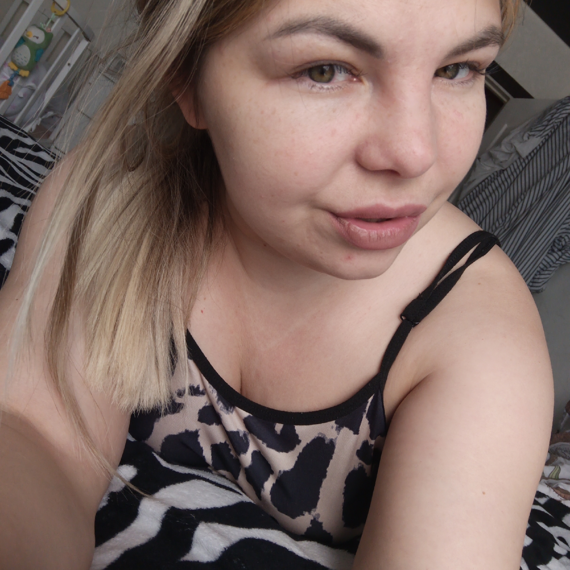A close-up, eye-level shot features a woman from the chest up, looking directly towards the camera. She has blonde or light brown hair that falls over her shoulders, and distinct brown or hazel eyes. Her expression is neutral to slightly pensive, with her lips subtly parted and adorned with light pink or nude lipstick. Freckles are visible across her cheeks and nose.
She is wearing a camisole with a black and beige/tan leopard print pattern and thin black spaghetti straps. The foreground is dominated by a black and white zebra-print blanket or sheet. In the background to the left, a white crib is visible, from which an owl-shaped mobile toy hangs. To the right, another patterned fabric, possibly a striped sheet, can be seen. The lighting suggests it is daytime, indicating an indoor setting, likely a bedroom or nursery due to the presence of the crib. No visible text is present in the image.