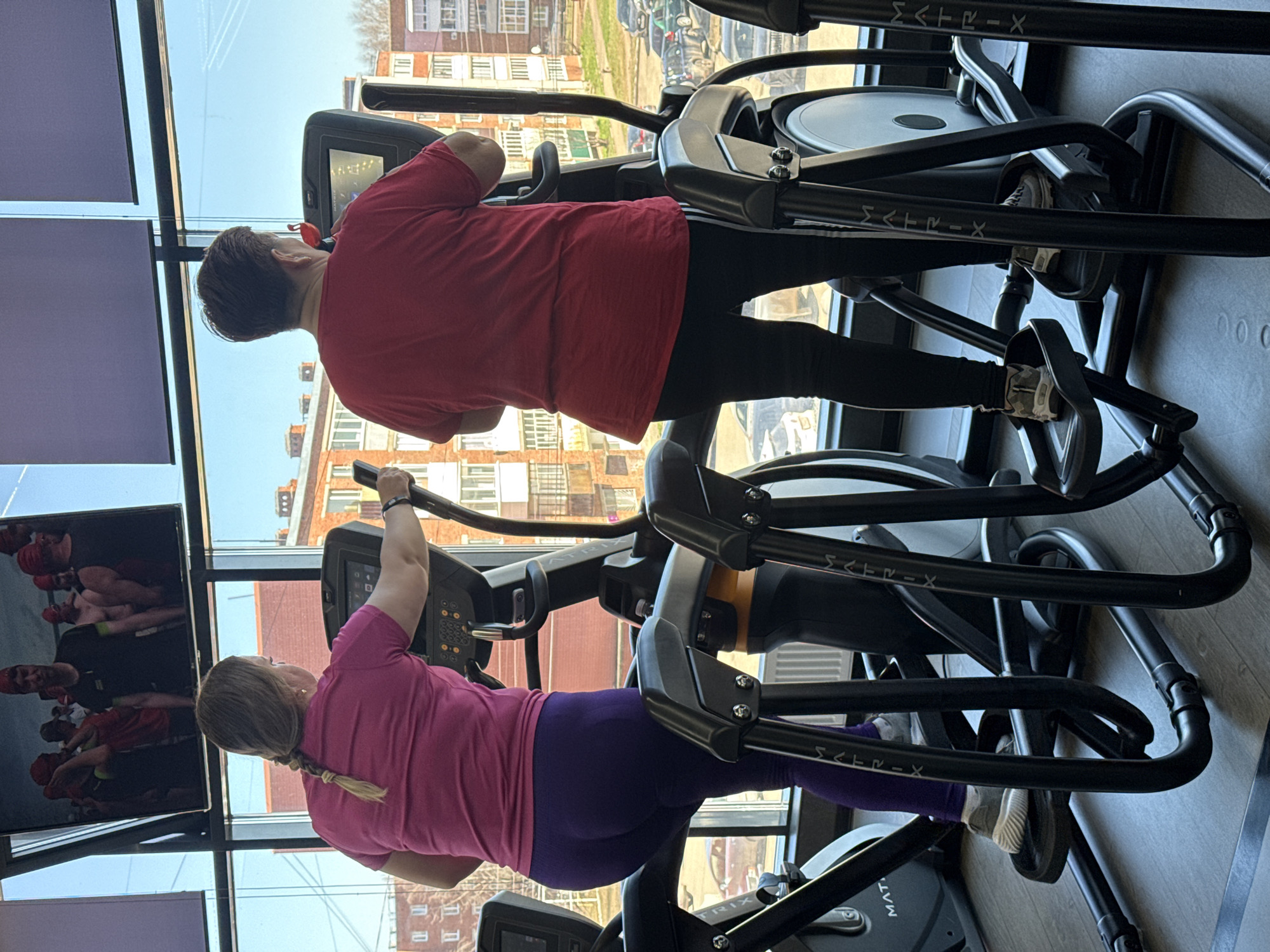 Two women are exercising on elliptical machines in a gym. The woman in the foreground is wearing a pink t-shirt and purple leggings and has her hair in a blonde braid. The woman behind her is wearing a red t-shirt and black leggings. Both are facing forward and holding onto the handles of their respective machines. Outside the window is a view of buildings, indicating an urban setting. The equipment has the brand name "MATRIX" on it. The gym appears to be in a building in Votkinsk, Russia. A television screen is visible on the left.