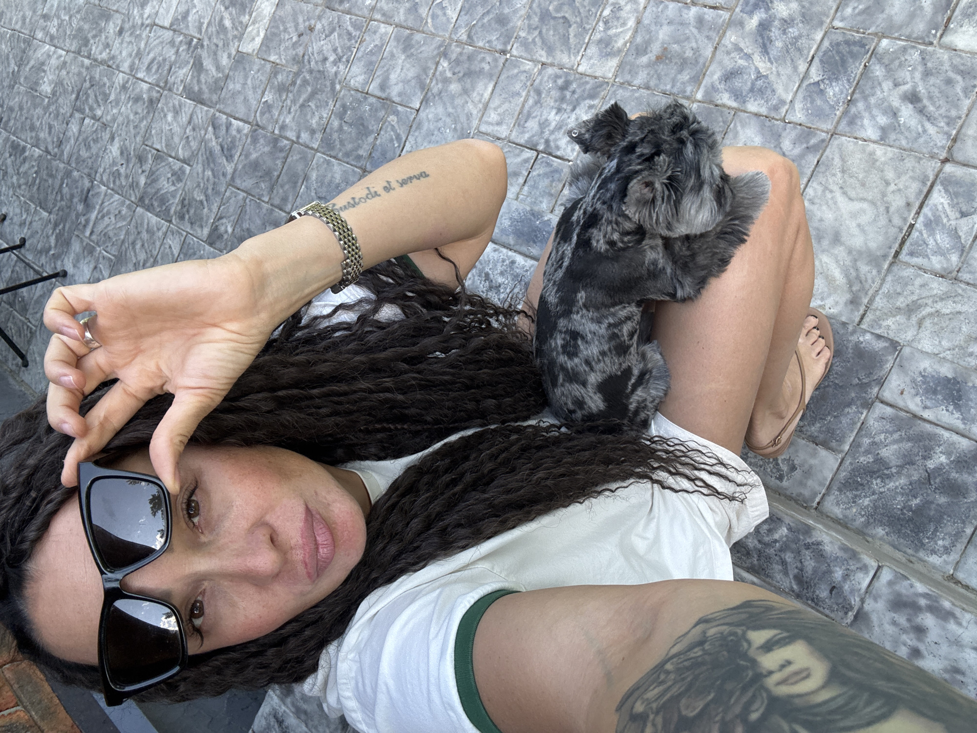A female subject is shown in a crouched position outdoors on a grey tiled surface in Chaikovsky, Russia. She has long, dark, curly hair and is wearing a light-colored t-shirt with a green trim on the sleeve. Her right hand holds black sunglasses slightly off her face, revealing her brown eyes. A large, detailed tattoo is visible on her left upper arm, and the phrase "custodi et serva" is tattooed on her right forearm, which also displays a metallic bracelet. A small, dark grey and black merle-patterned dog rests on her lap. Her right foot, wearing a light-colored flat shoe, is partially visible on the ground. The sunglasses reflect an outdoor scene with clouds and trees. The environment consists of textured, grey rectangular pavers.