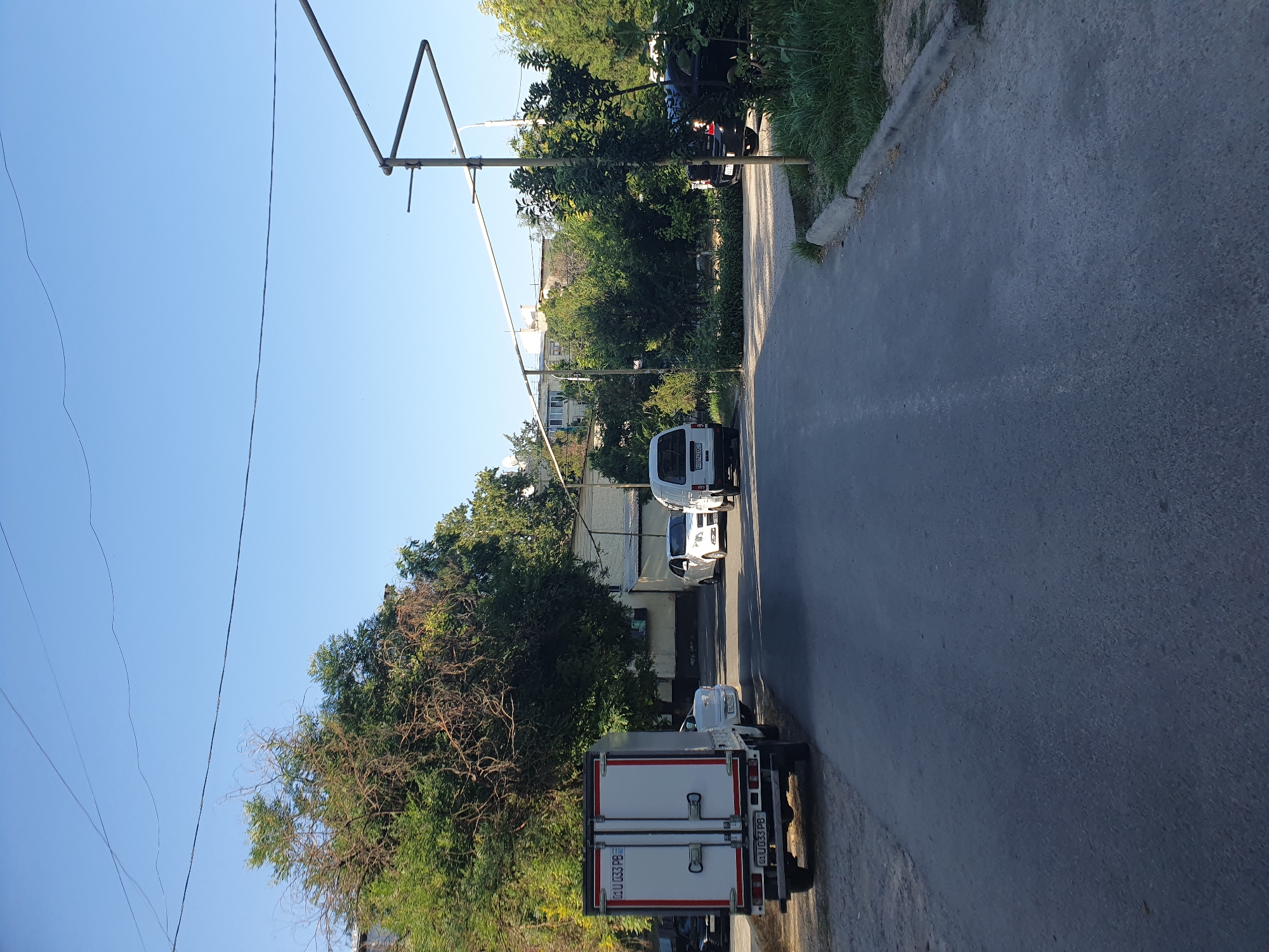 The image captures a street scene in Tashkent, Uzbekistan, under a clear blue sky. In the foreground, a gray asphalt road stretches into the distance, bordered by a concrete curb and overgrown grass on the right. On the left side of the road, a white box truck with red trim is parked, displaying license plates that read "01 U 033 PB". Further down the street, a white sedan and a white van are visible, also parked. Behind the vehicles, buildings and mature trees with green and brown foliage line the street. A metal structure, possibly a canopy frame, is present in the mid-ground. The overall impression is a typical urban residential street.