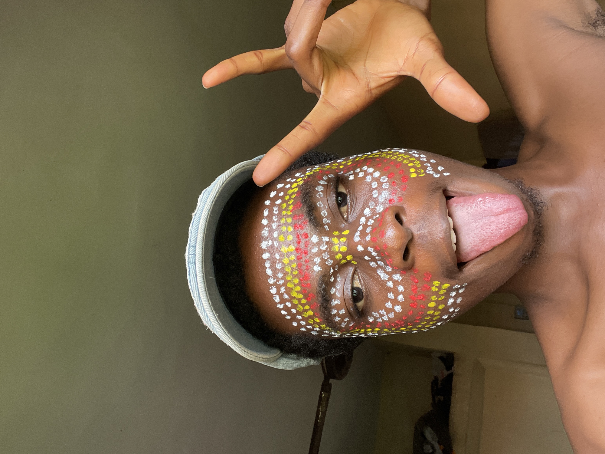 A man with painted face, displaying red, white, and yellow dots, is sticking his tongue out, in an indoor setting, possibly Nkpor, Nigeria. He is wearing a hat and making a hand gesture. The background is a wall with a door.