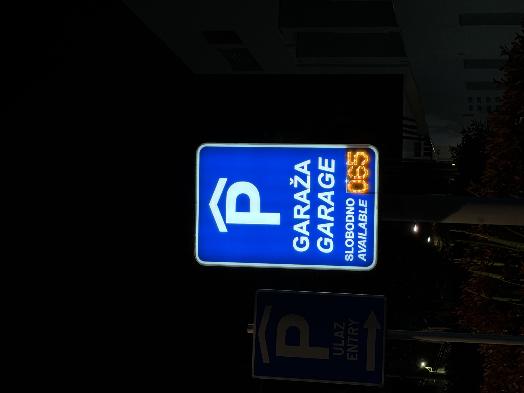 The image shows two illuminated parking garage signs at night. The top sign is larger and displays a white "P" symbol, the words "GARAŽA GARAGE", "SLOBODNO AVAILABLE", and a digital display showing "065". The sign is illuminated in blue and white. Below this sign is a smaller blue sign with a white "P" symbol, the words "ULAZ ENTRY", and an arrow pointing to the right. The background is dark, with some faint building details and foliage visible. The location is Trebesin, Montenegro.