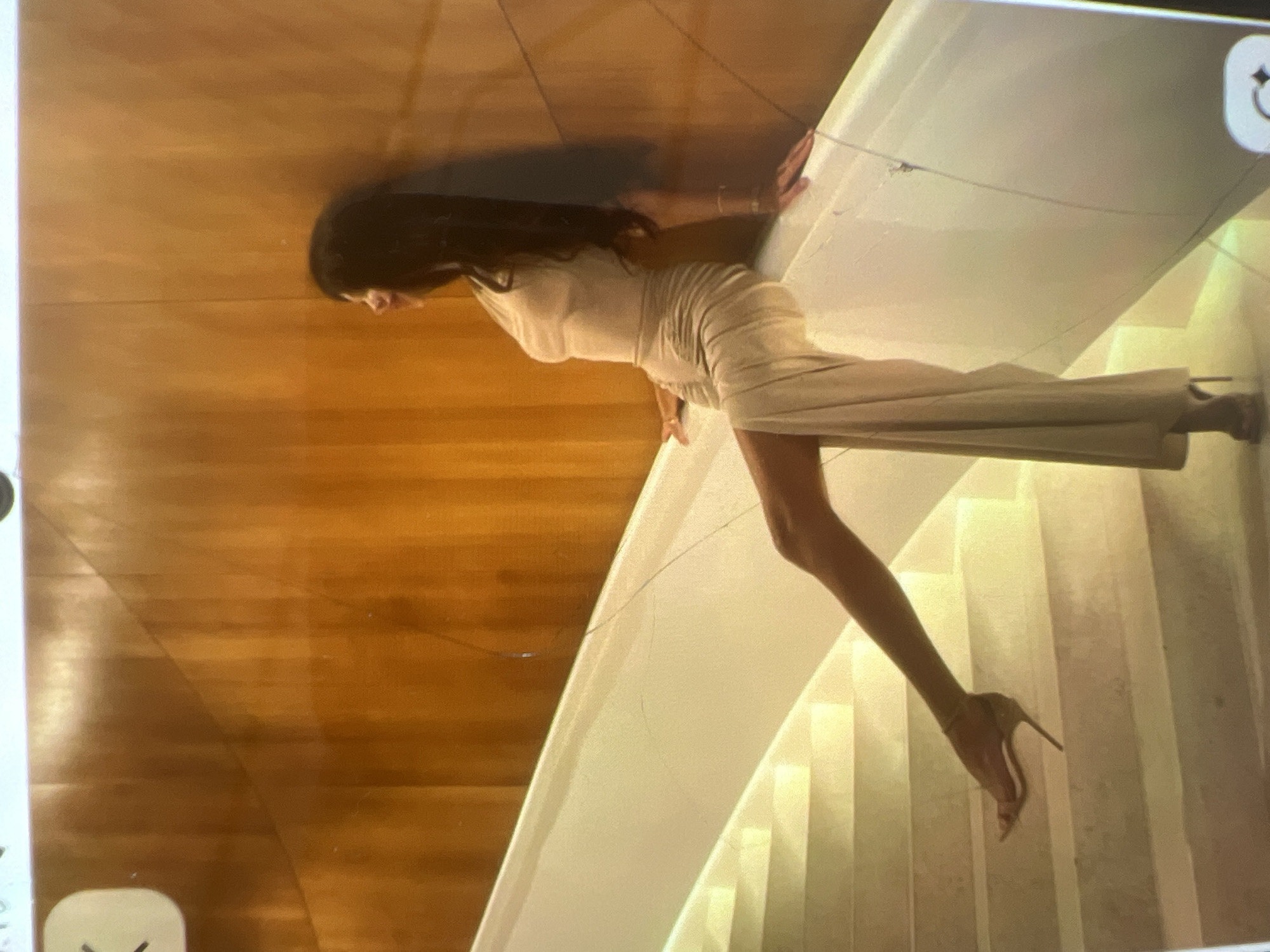A woman with long dark hair is sitting on the edge of a white staircase, leaning against a wood-paneled wall. She is wearing a light-colored, form-fitting dress with a high slit up the leg, and high-heeled sandals. Her legs are extended, with one foot resting on a lower step of the stairs. The wall behind her has vertical wood paneling. The scene appears to be indoors, possibly a modern building. There is no indication that this location is in the city of Kara-Dıykan, Kyrgyzstan.