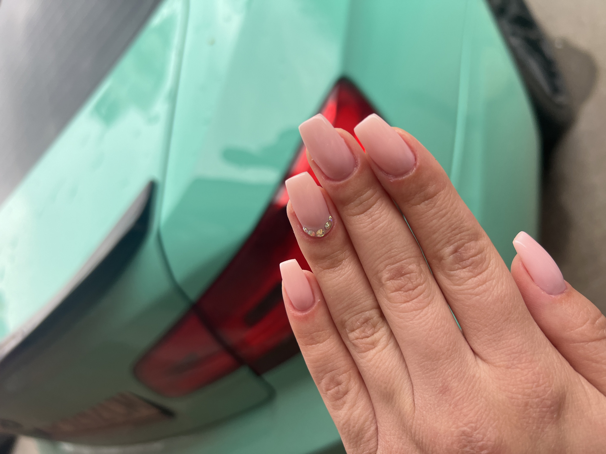 The image displays a close-up of a person's right hand showcasing a fresh manicure, set against a softly blurred background. The nails are long, shaped in a coffin or ballerina style, and painted in a natural light pink or nude shade. The ring finger's nail is adorned with a small cluster of clear or iridescent rhinestones near the cuticle. The skin on the hand appears smooth and well-kept.
In the background, the distinct, vibrant teal or light green paintwork of a vehicle is visible, likely the rear bumper or trunk area. A prominent red, rectangular taillight is also partially visible on the right side of the frame. The lighting suggests an outdoor, daytime setting. The main activity depicted is the presentation of the manicured nails. There is no visible text in the image. The contextual location for this scene is Plovdiv, Bulgaria.