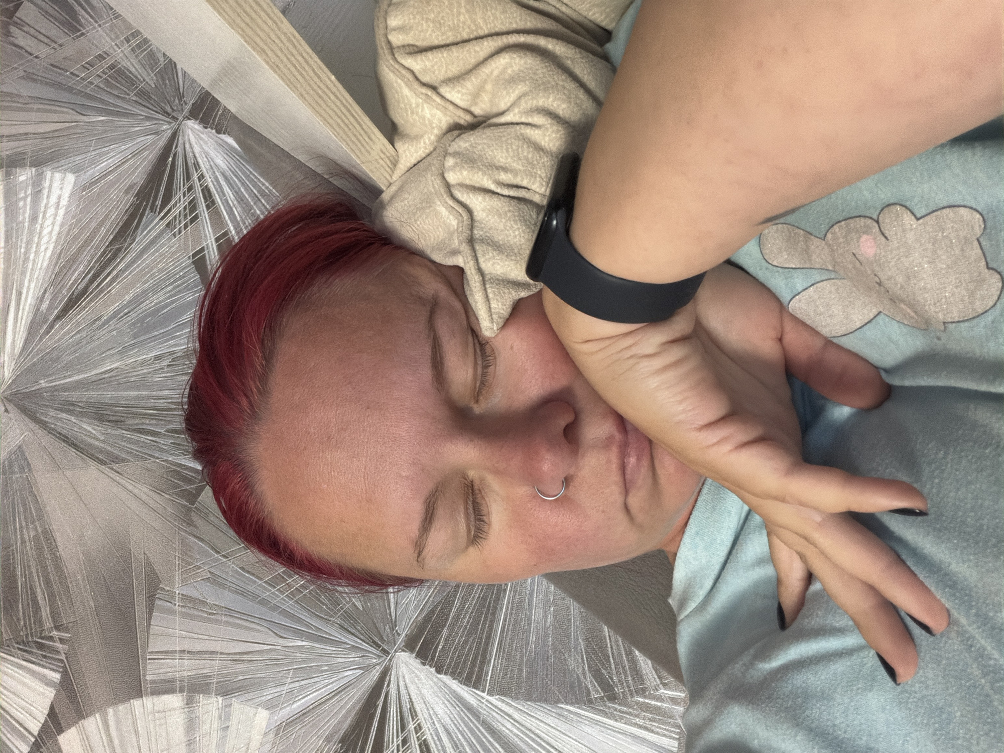 The image shows a close-up of a person with red hair sleeping, resting their head on their arm. The person is wearing a light blue t-shirt with a bunny graphic and a beige long-sleeved shirt. A dark smartwatch is visible on their left wrist. The background consists of a patterned wallpaper. There is no indication of location in the city of Chaikovsky, Russia.