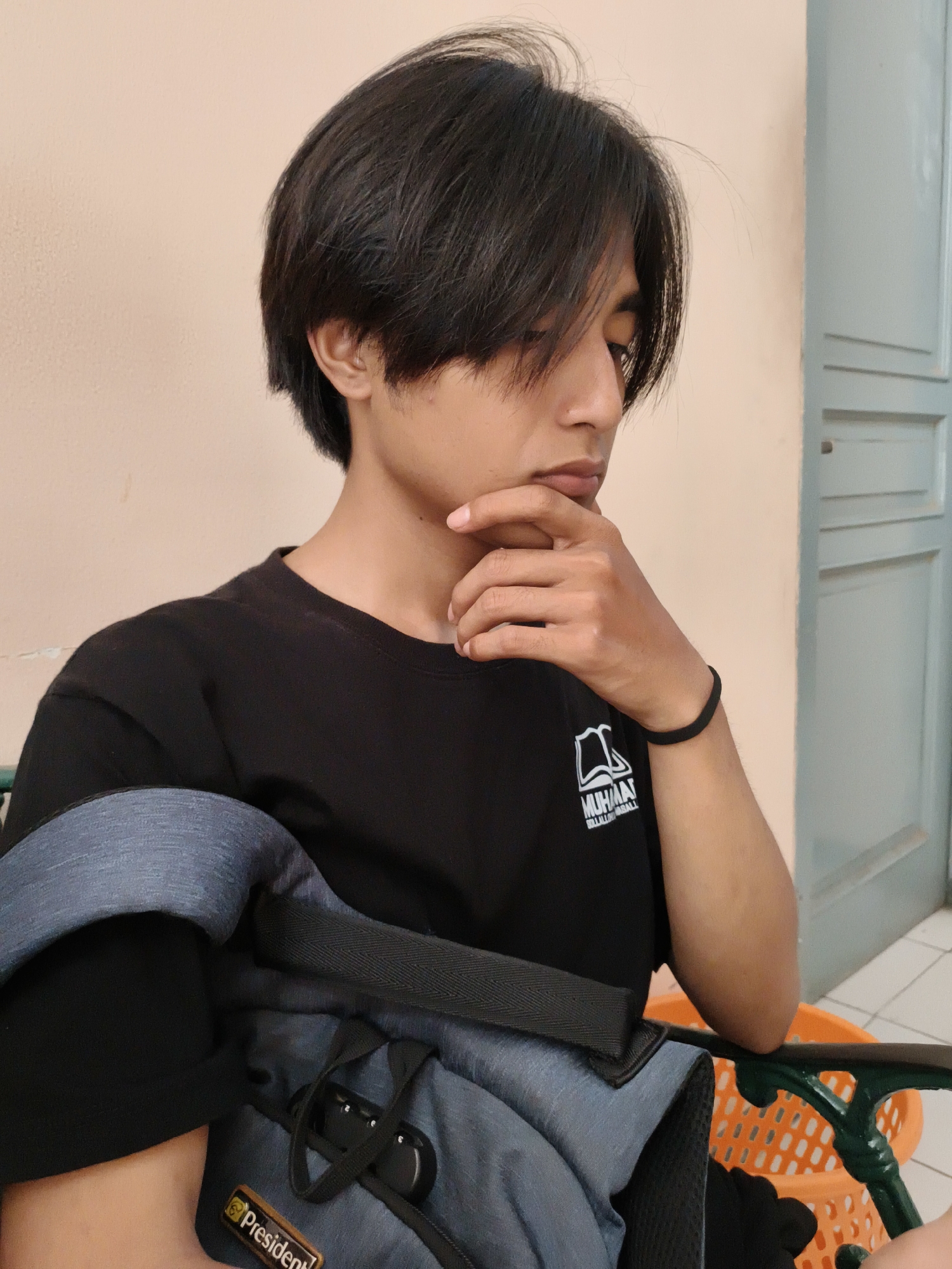 A young man with black hair is seated, appearing thoughtful. He is wearing a black t-shirt with a white graphic on the left side. He rests his right hand on his chin. A blue and black backpack is in his lap, branded with a "President" label. The background consists of a light peach wall and a light blue door. The location is likely in Cianjur, Indonesia.