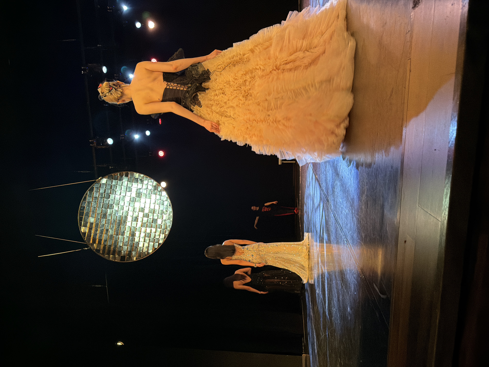 The image depicts a fashion show in Blagoevgrad, Bulgaria. A woman in a large, ruffled, peach-colored gown and black corset is standing on a stage. Below her, another woman in a shimmering silver dress is also on the stage, along with a third woman in a black dress. In the background, there is a large, circular disco ball. Stage lighting is visible. There is another figure in the background. The stage appears to be made of wood.