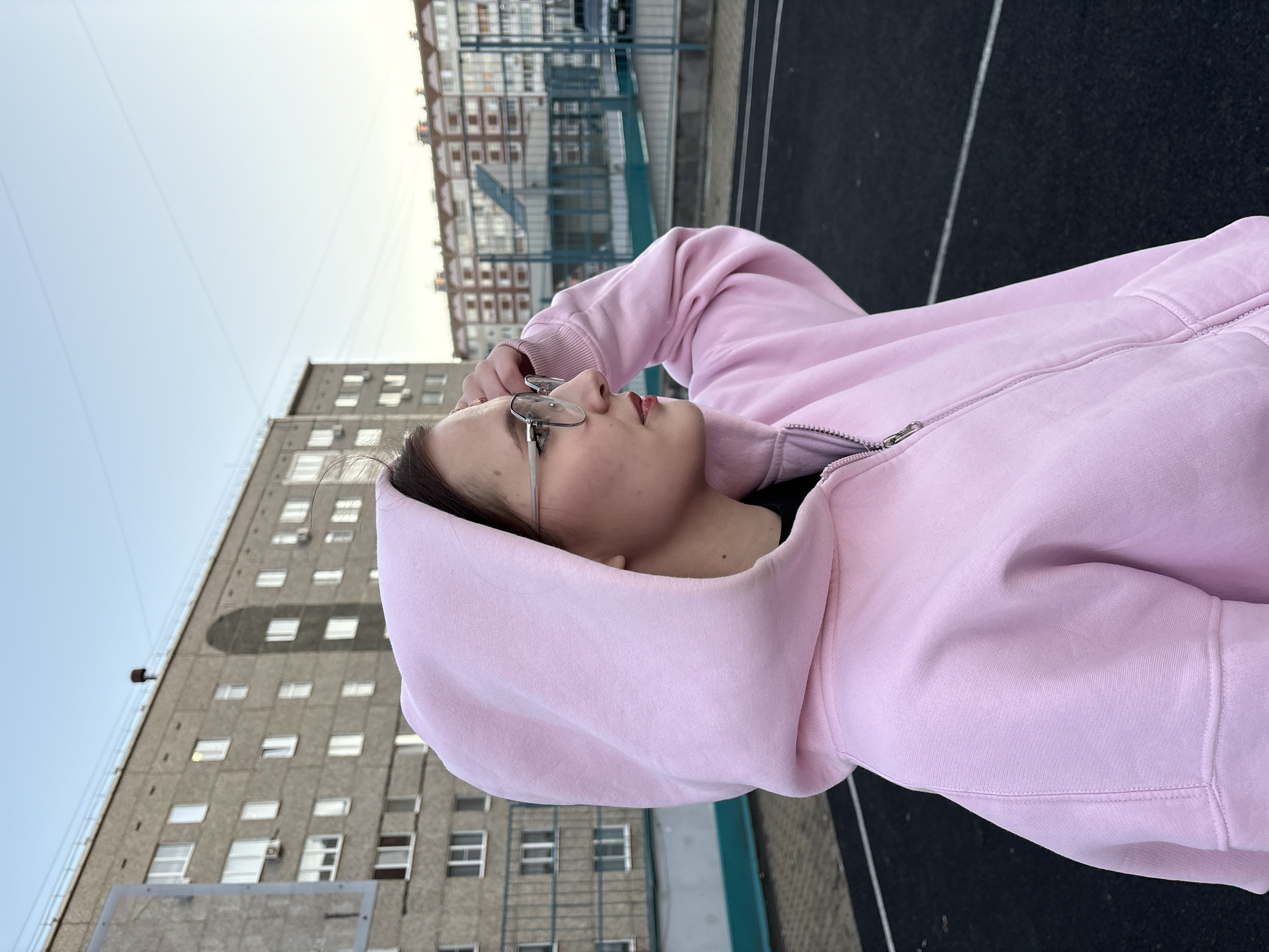 A young woman is depicted in the photograph, wearing glasses and a pink hooded sweatshirt. She is standing on a black surface, which appears to be a basketball court. Behind her are buildings typical of urban architecture in Izhevsk, Russia. The sky is visible in the background, suggesting it is daytime. She is looking slightly upward with her hand near her glasses.