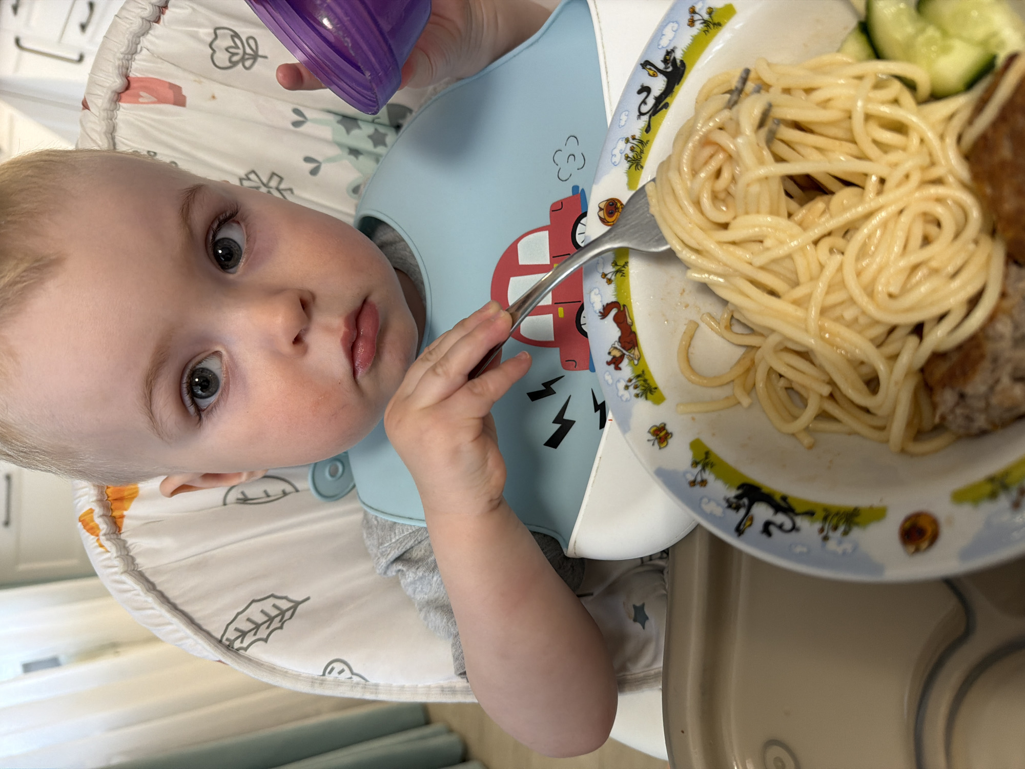 A young child with blonde hair and grey eyes is sitting in a highchair, wearing a light blue bib with a red car design. The child is holding a purple cup and a fork, with spaghetti, a meatball, and sliced cucumbers on a plate in front of them. The plate has a cartoon animal design. The highchair has a white patterned seat cover. The scene is indoors, with a glimpse of a white cabinet in the background. The location is not discernible as Votkinsk, Russia from the provided image.